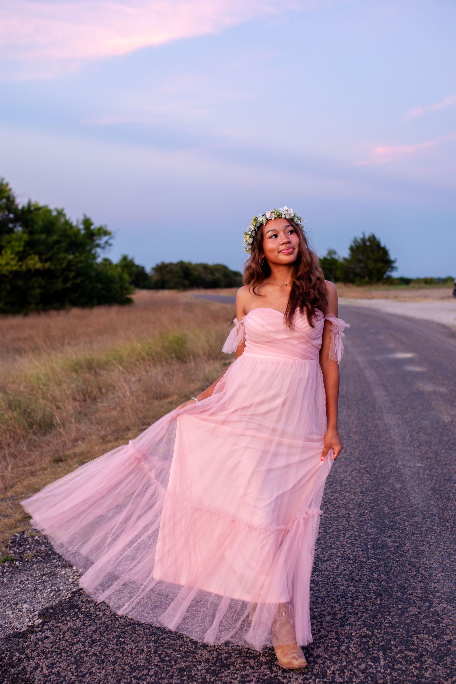 A figure in a flowing pink tulle dress twirls gracefully on a rural road during sunset creating a dreamy atmosphere.