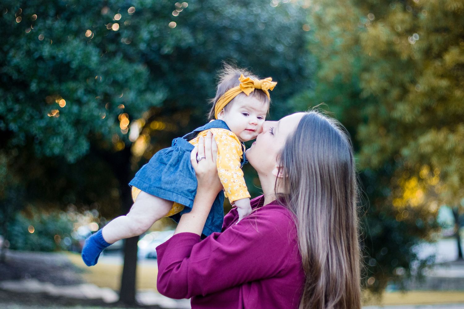 A caregiver lifts up a small child wearing blue overalls outdoors during a golden sunset.