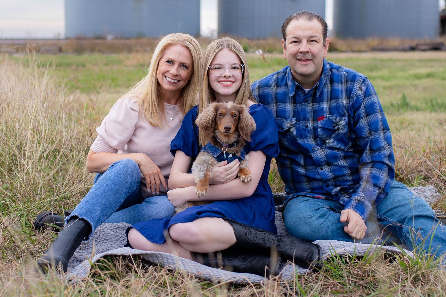 Family relaxes together on blanket in grassy field during outdoor photo session at sunset.