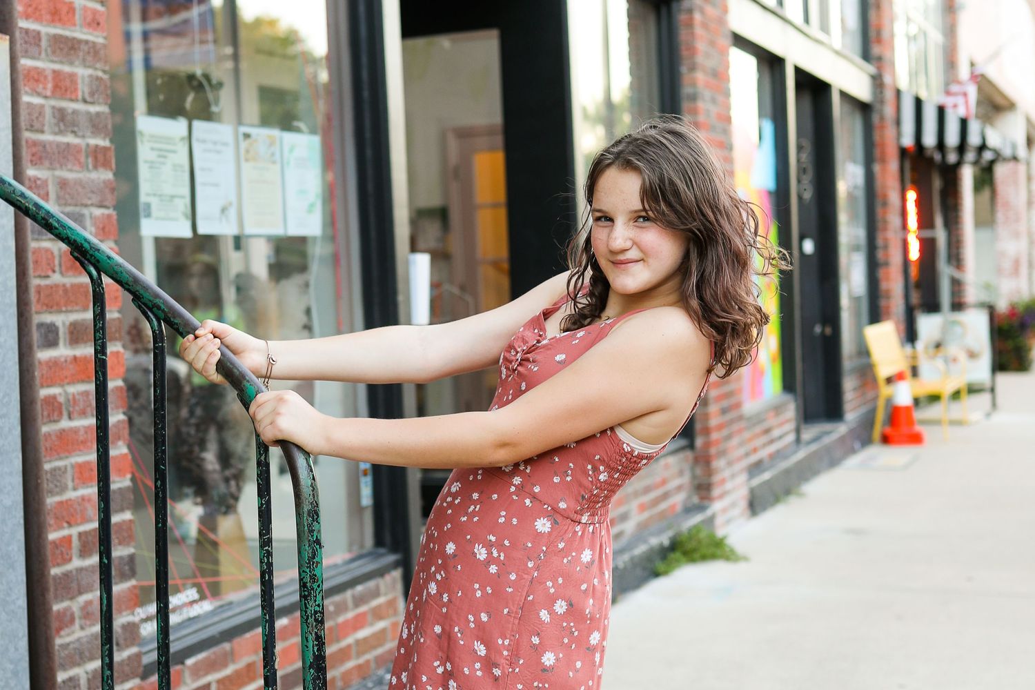Woman in floral dress leans against railing on urban sidewalk with brick buildings in background.