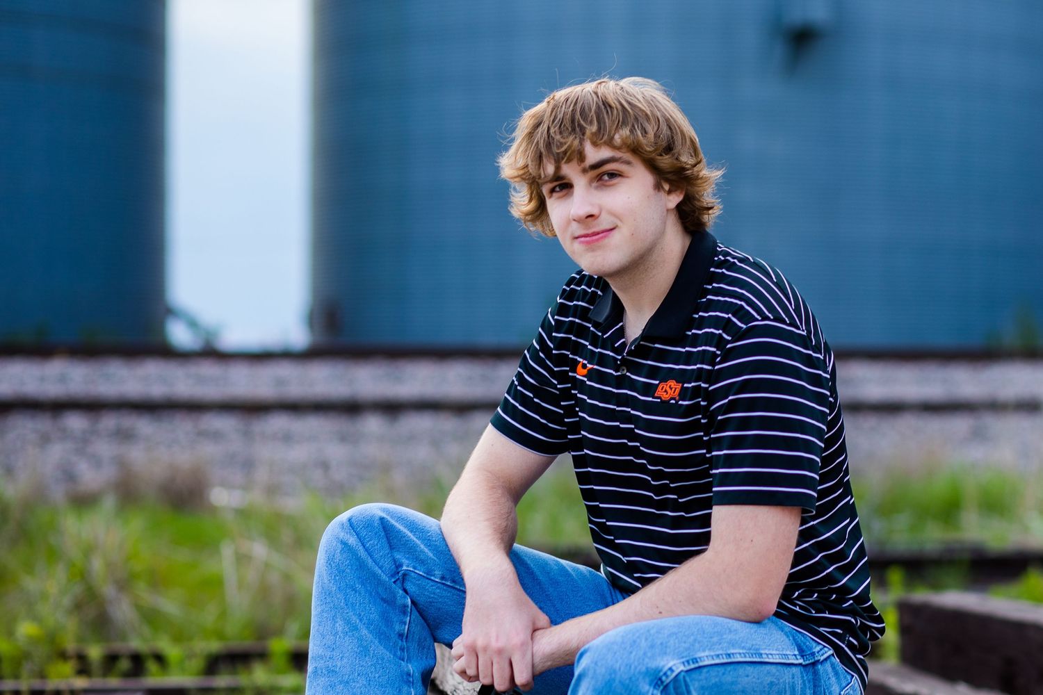 Person wearing striped polo shirt and blue jeans sitting casually outdoors against blue wall background.
