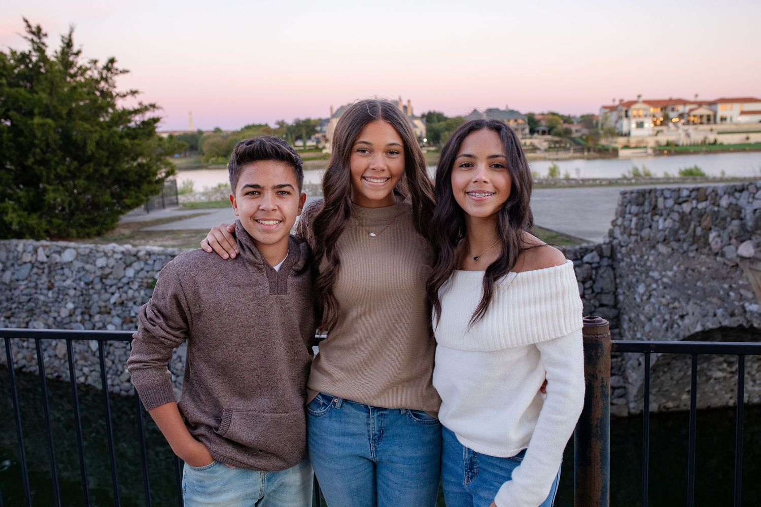 Three young people smiling and posing together outdoors at sunset with houses and stone walls in background.