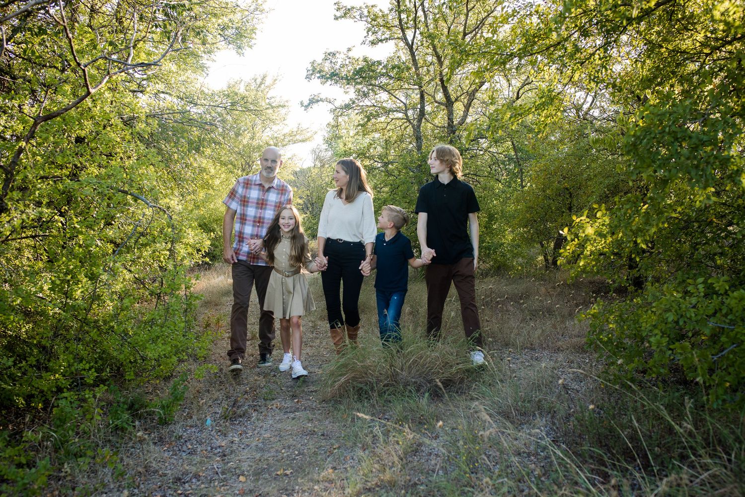 Group of people walking together on a sunlit nature trail surrounded by green trees and foliage on a bright day.