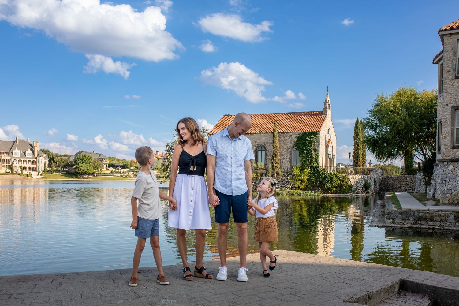 Family stands together on a lakeside path with beautiful houses and church in background on sunny summer day.