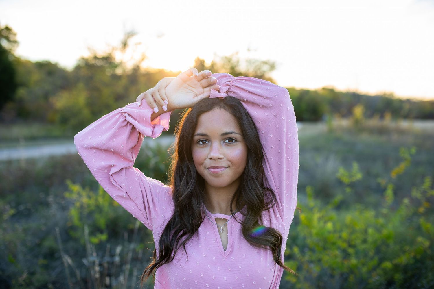 Person wearing pink sweater poses in golden evening sunlight among wildflowers during sunset photoshoot.