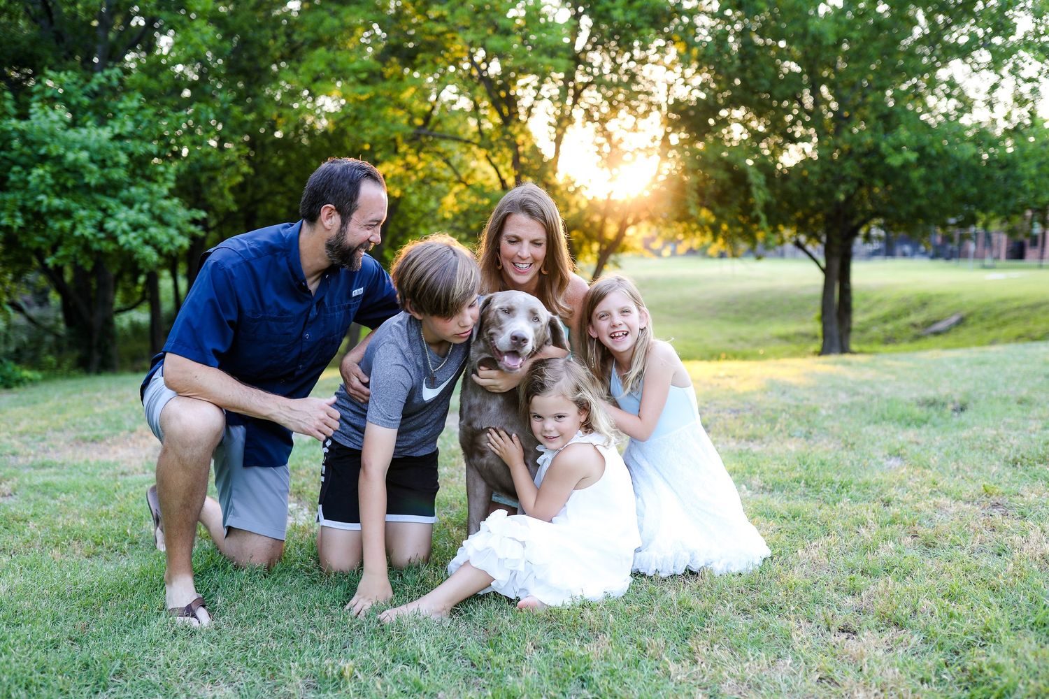 A loving group sits together on grass with their brown dog during golden hour sunset in a park setting.