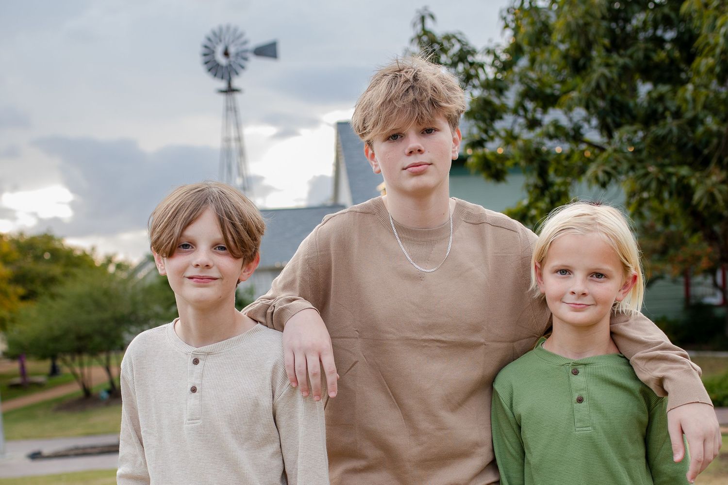 Three young sunlit figures stand close together outdoors in coordinated neutral-toned casual outfits.