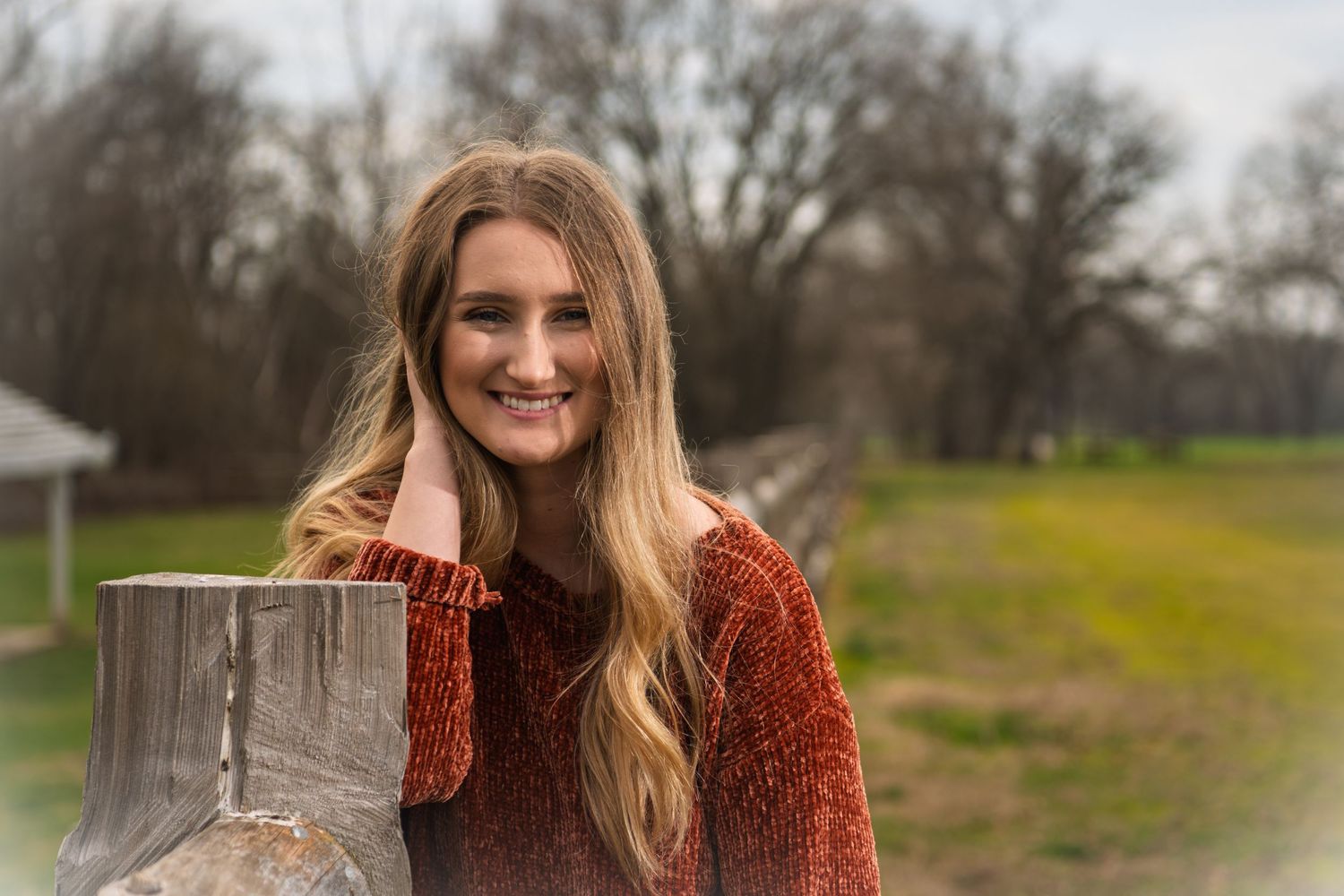 A person in a cozy rust-colored sweater smiles while leaning against a wooden fence post in a rural setting.