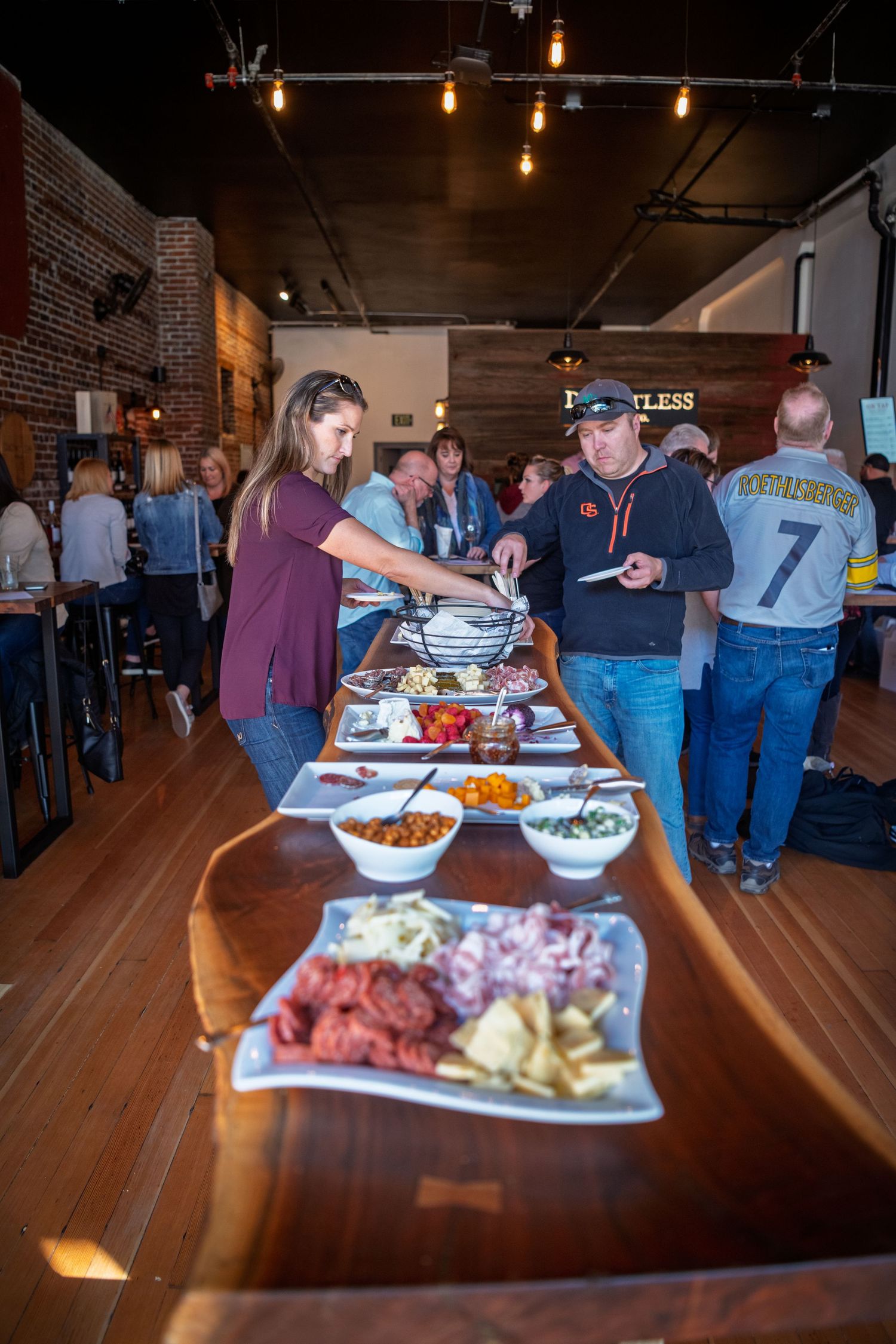 People gather around a buffet table with food and drinks at an indoor event with exposed brick walls and hanging lights.