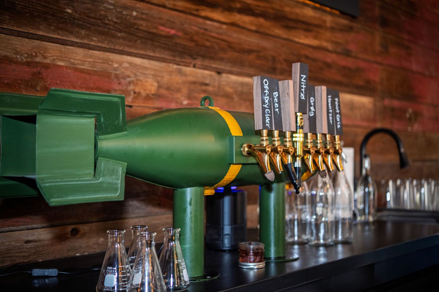 Wooden bar counter with rows of empty glass bottles and green tap handles in a rustic tavern setting.