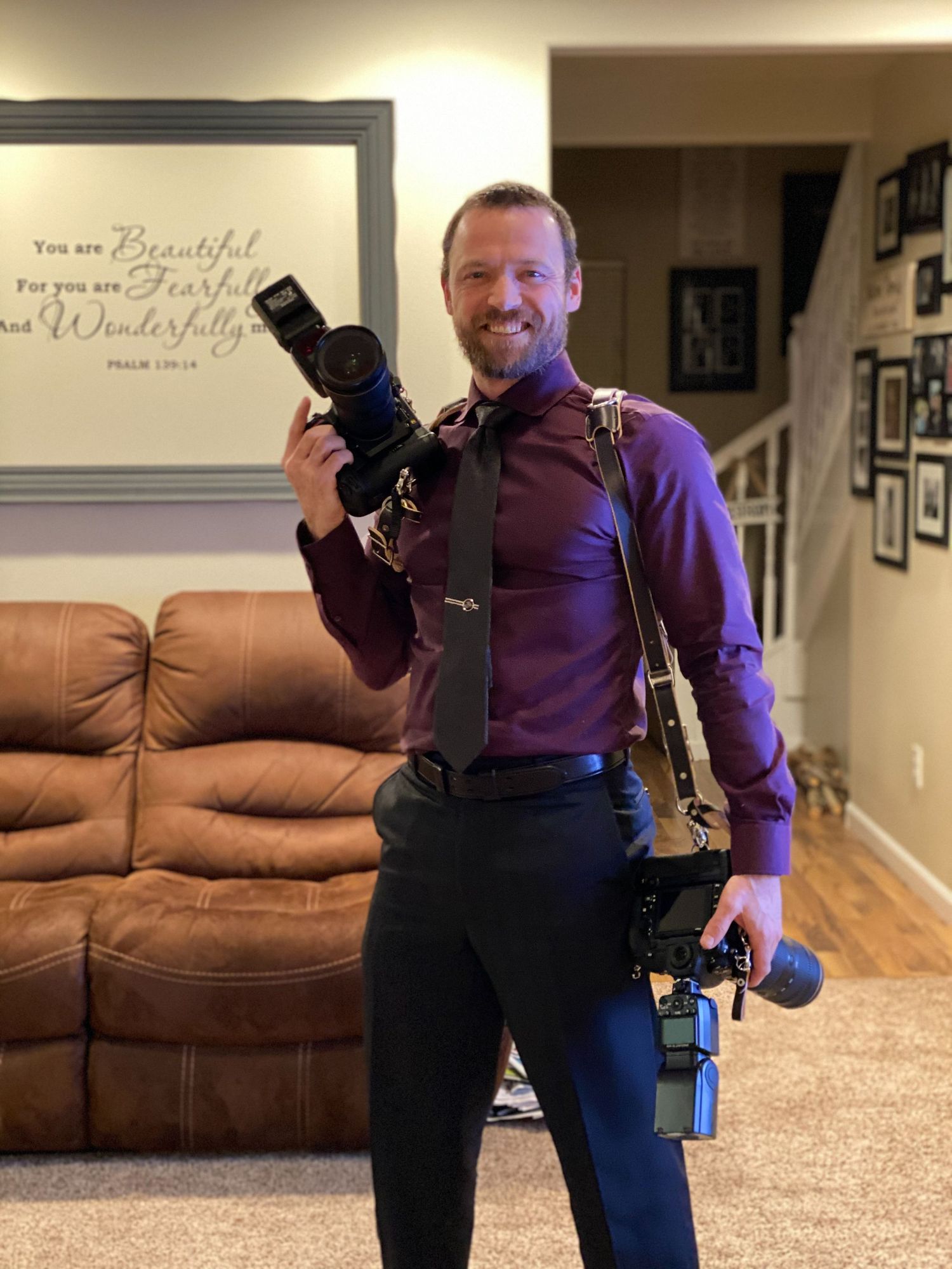 Professional photographer smiling with cameras in hand, wearing dress shirt and tie, ready for a photo session.