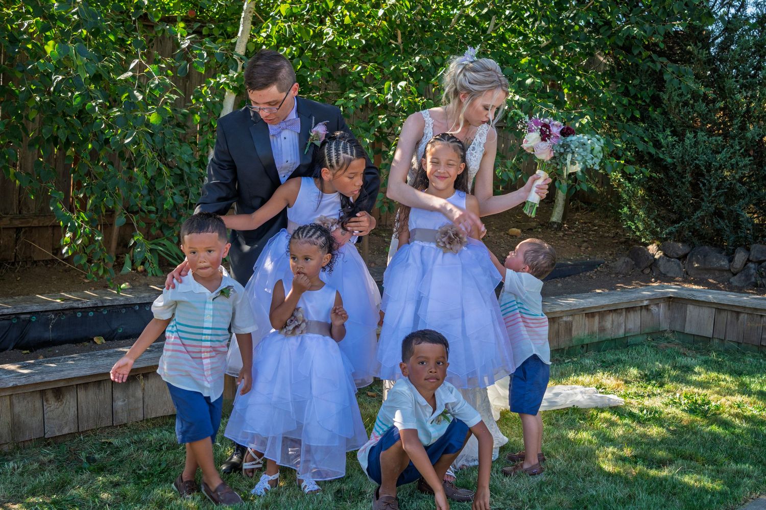 A group gathers outdoors in formal attire with children in white dresses and blue outfits during a sunny garden celebration.