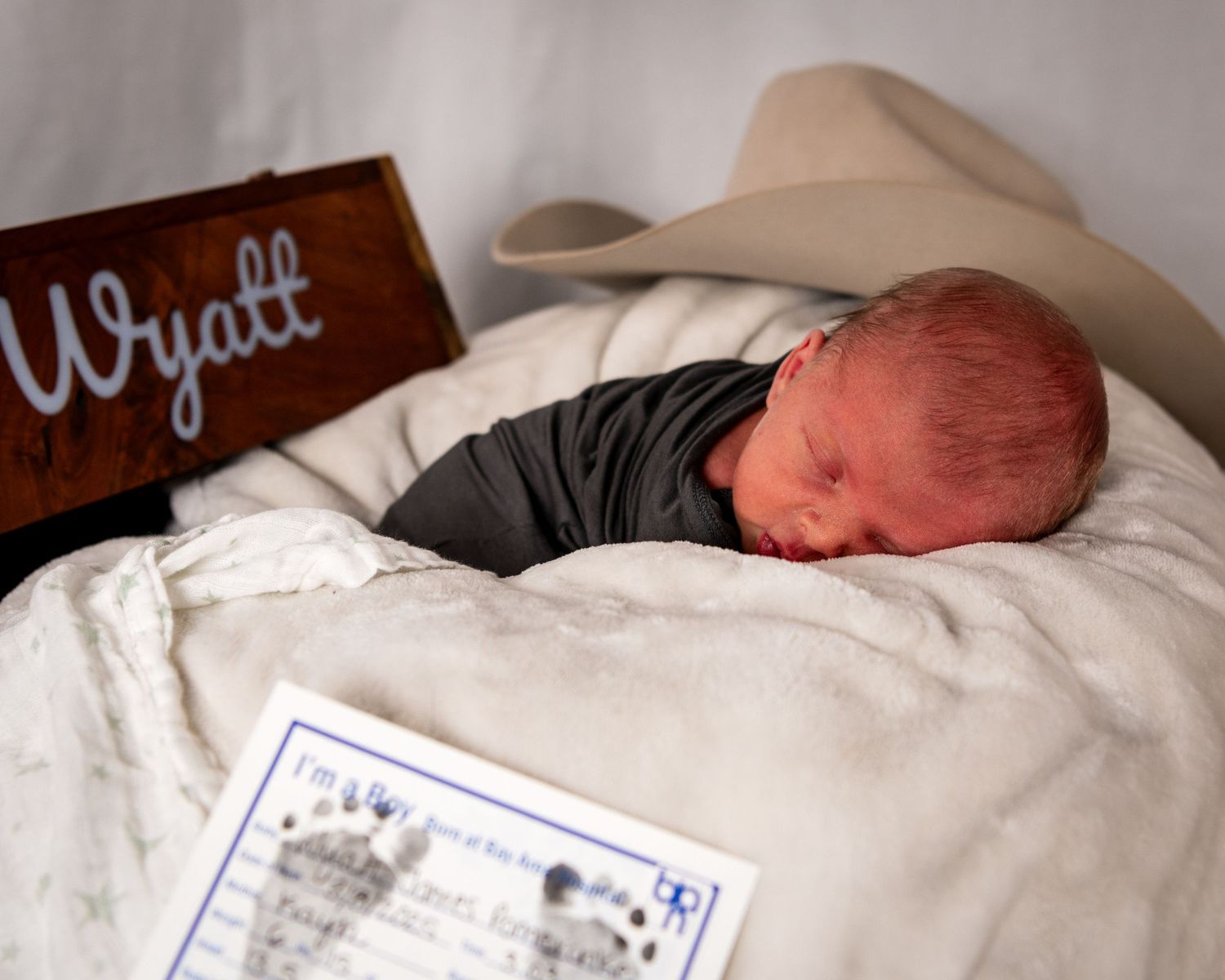 Rustic newborn portrait with baby sleeping under cowboy hat, personalized name sign, and soft blanket.