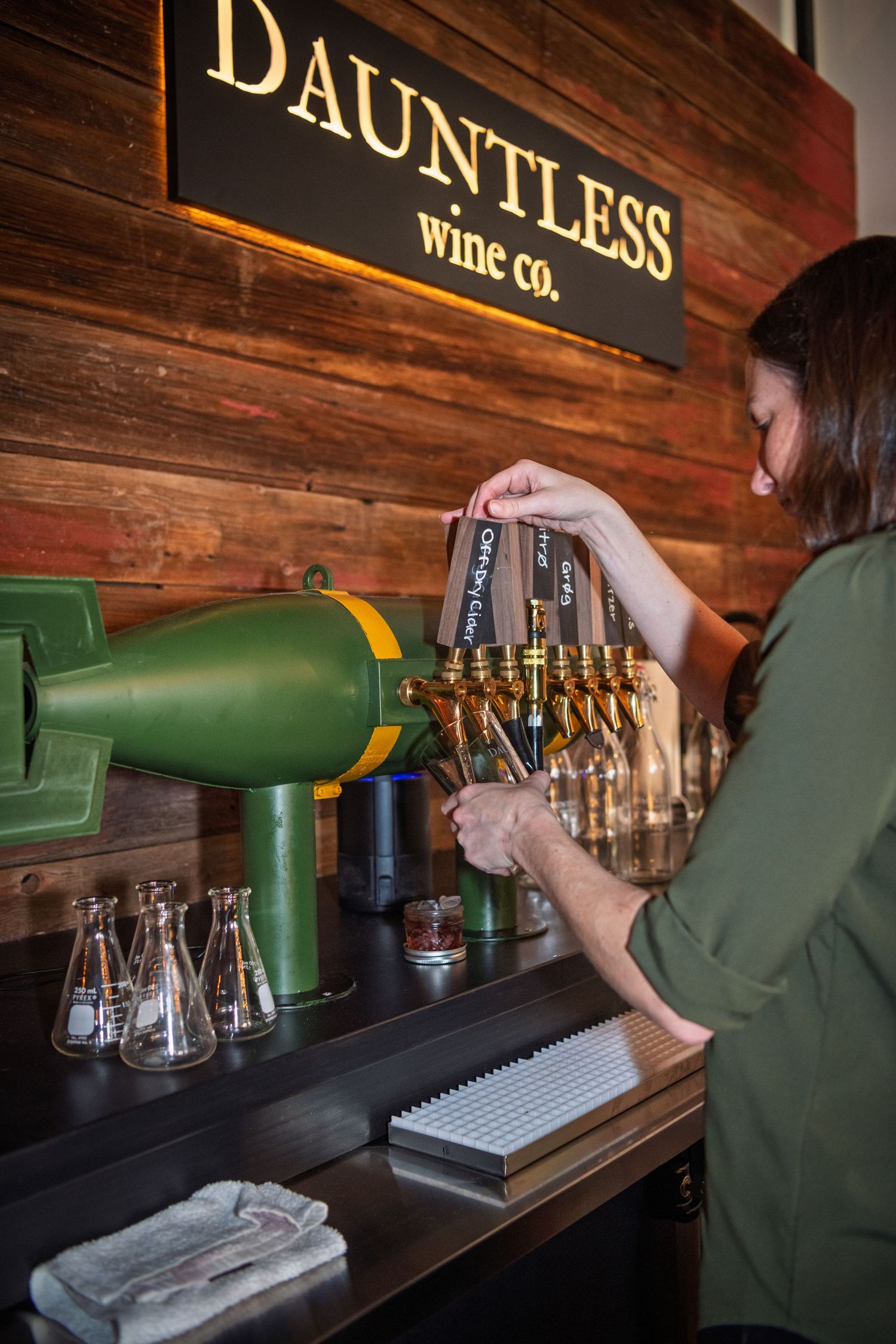 Hands pour beer from green taps at a rustic wooden bar with a stainless steel counter and glasses lined up.