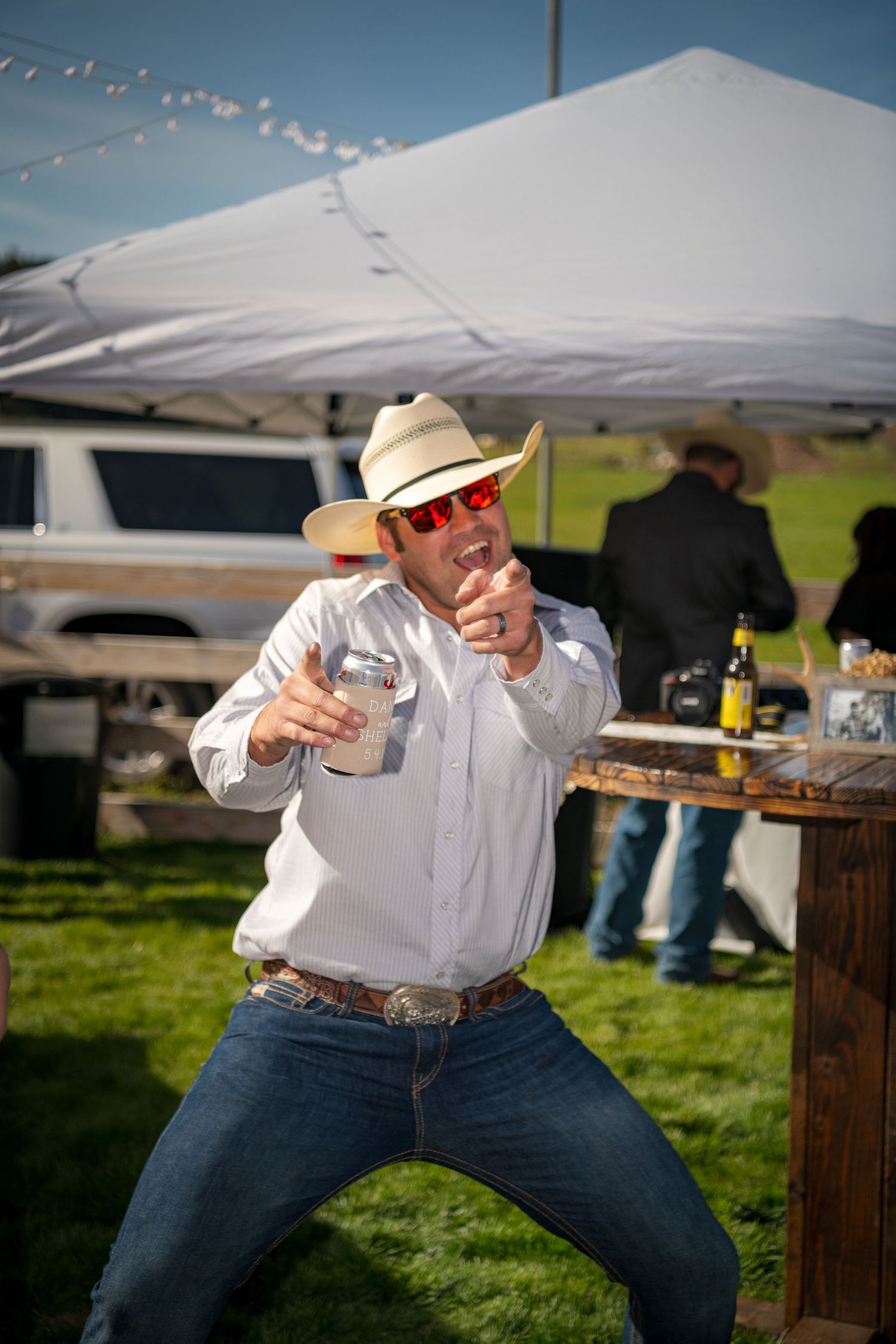 Candid wedding photography capturing cowboy guest celebrating with laughter and drinks at outdoor reception.