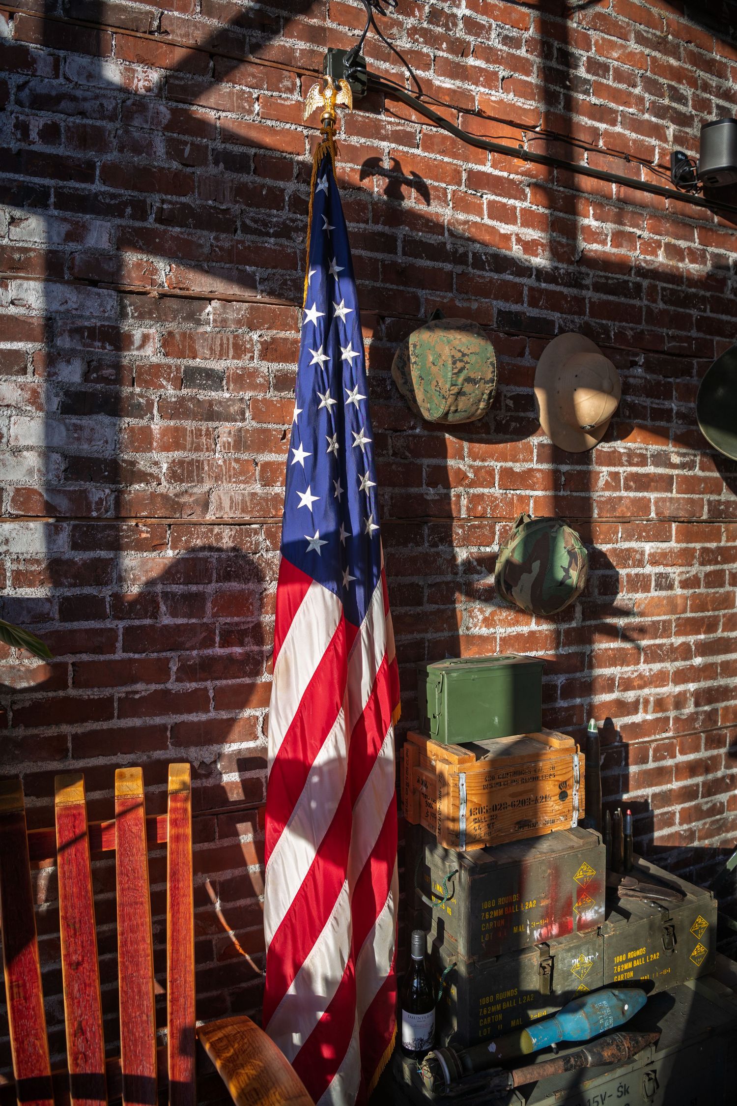American flag displayed against rustic brick wall with vintage military equipment and wooden crates in warm sunlight.