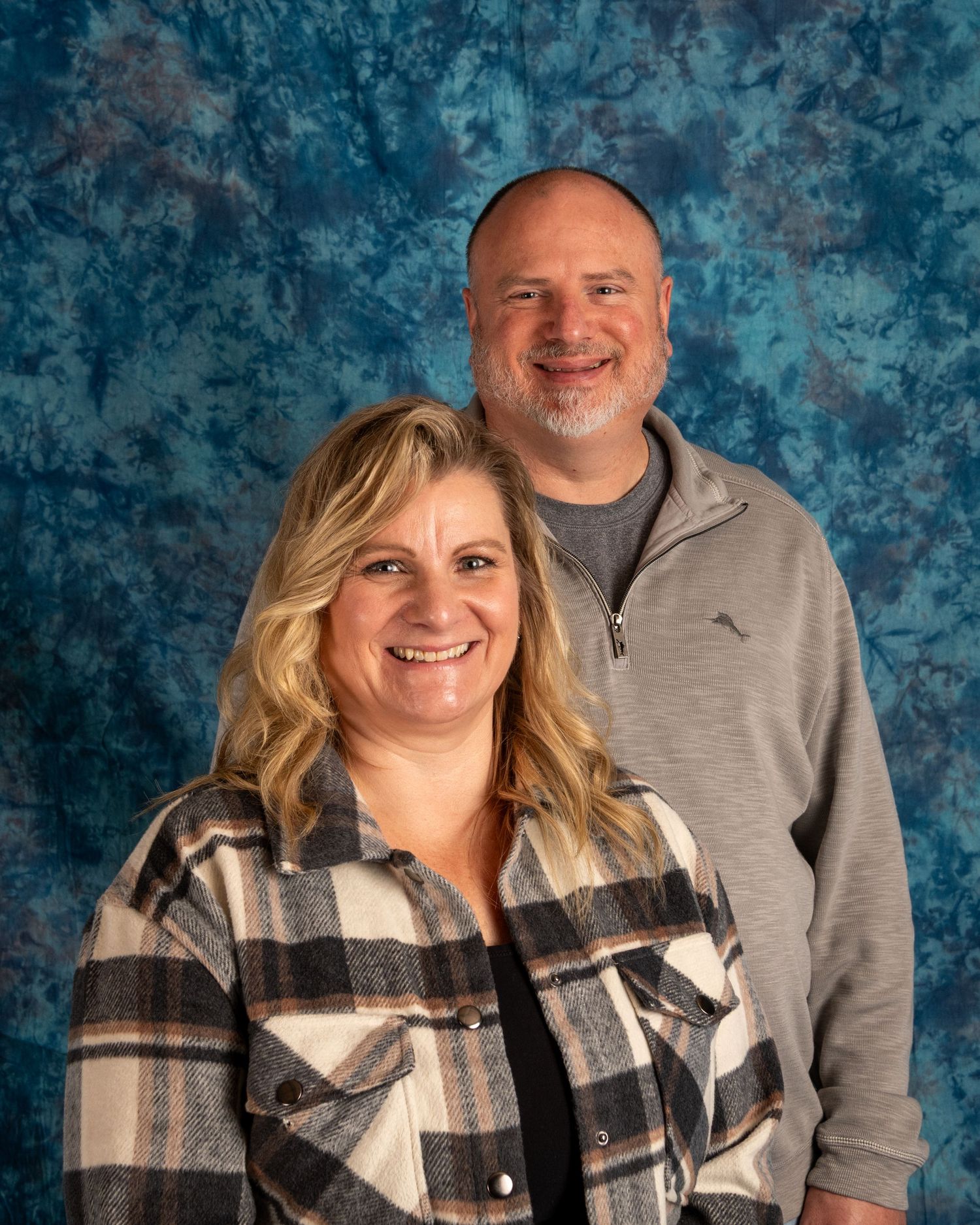 A couple poses together for a portrait against a mottled blue backdrop, wearing casual attire including a plaid shirt and grey pullover.