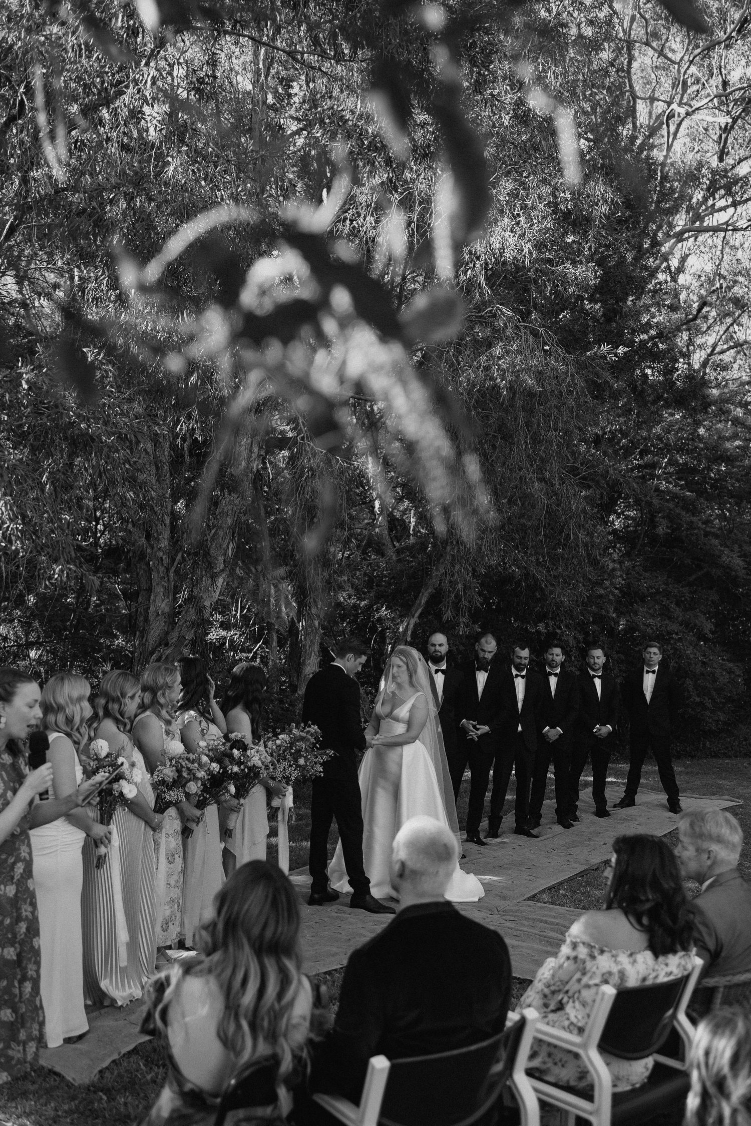 Black and white photo of outdoor wedding ceremony with bridal party and guests among tall trees.