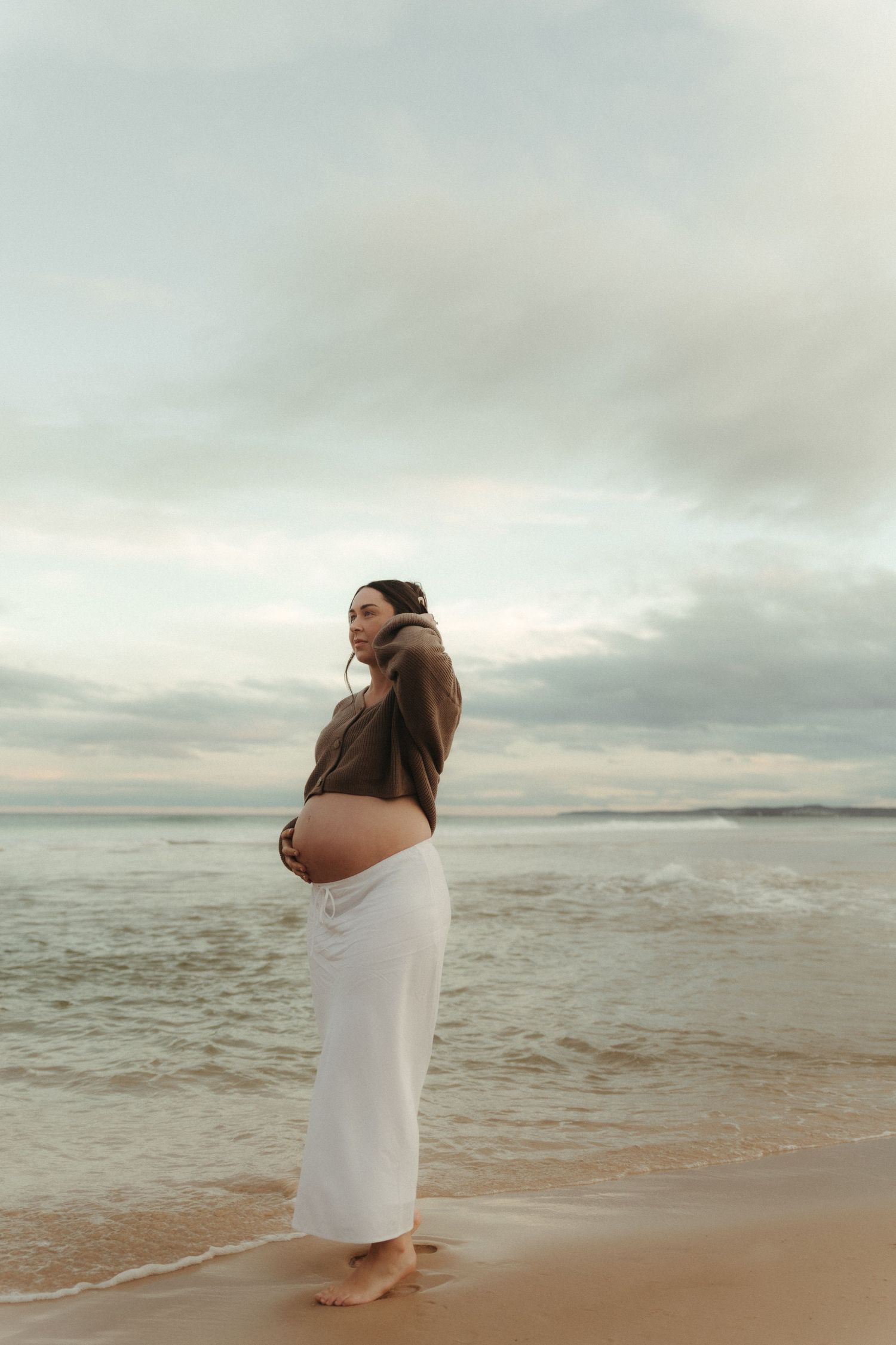 Pregnant woman in brown hoodie and white skirt cradling her baby bump on a serene beach at dusk.