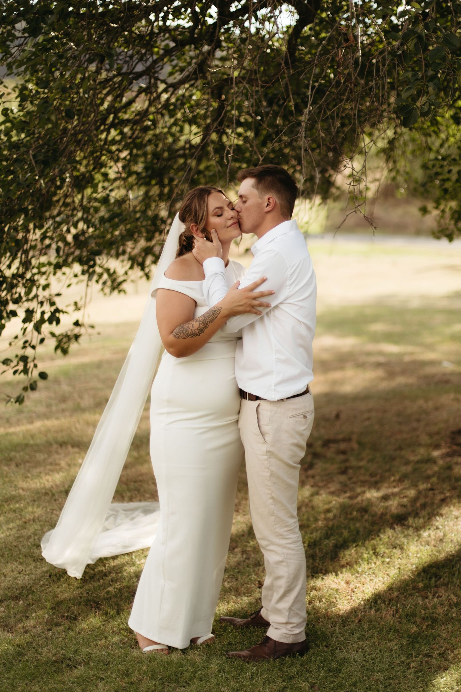 Bride and groom share a romantic kiss under a tree in outdoor wedding portraits.