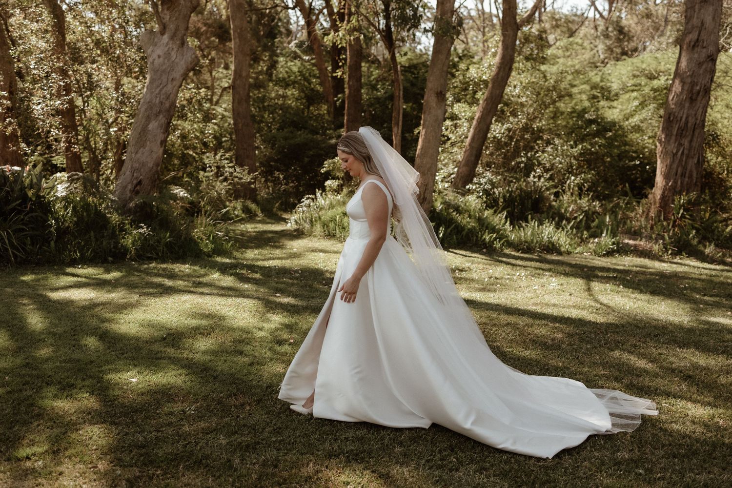 Elegant bride in a flowing white gown and veil posing in a lush green garden setting.