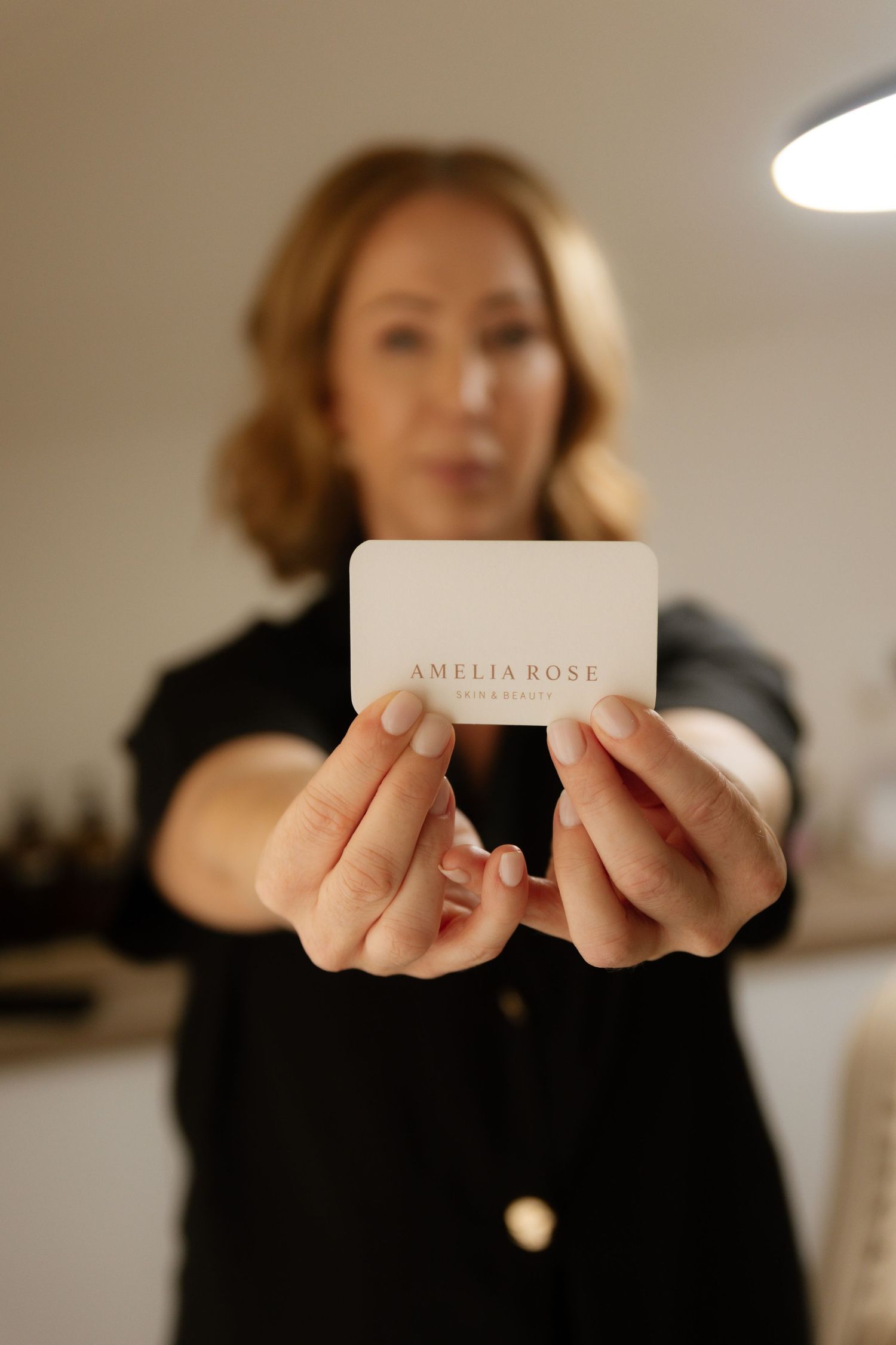 Woman holding Amelia Rose business card toward camera in a softly lit beauty studio.