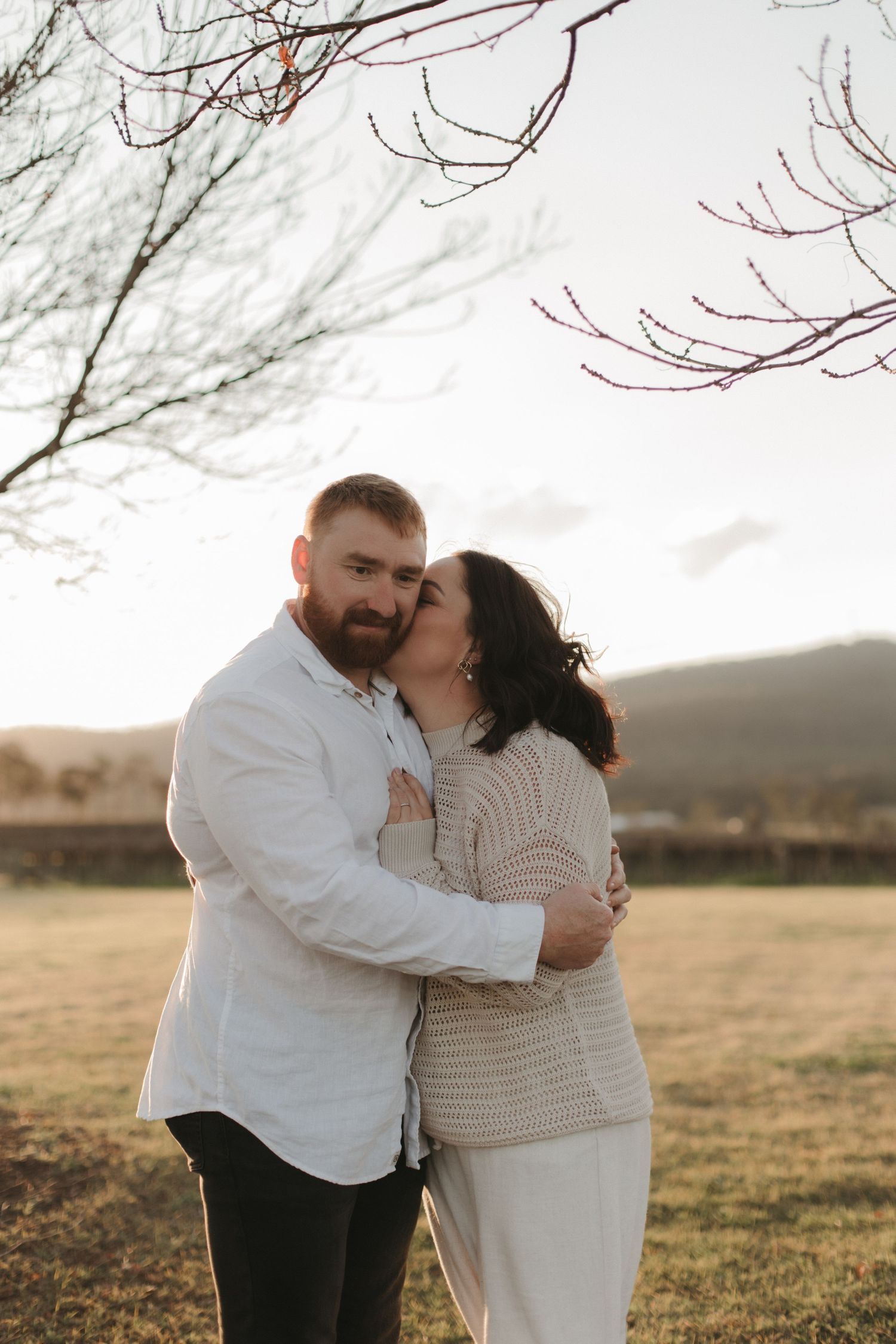 Couple embracing and kissing outdoors with bare tree branches and mountains behind.