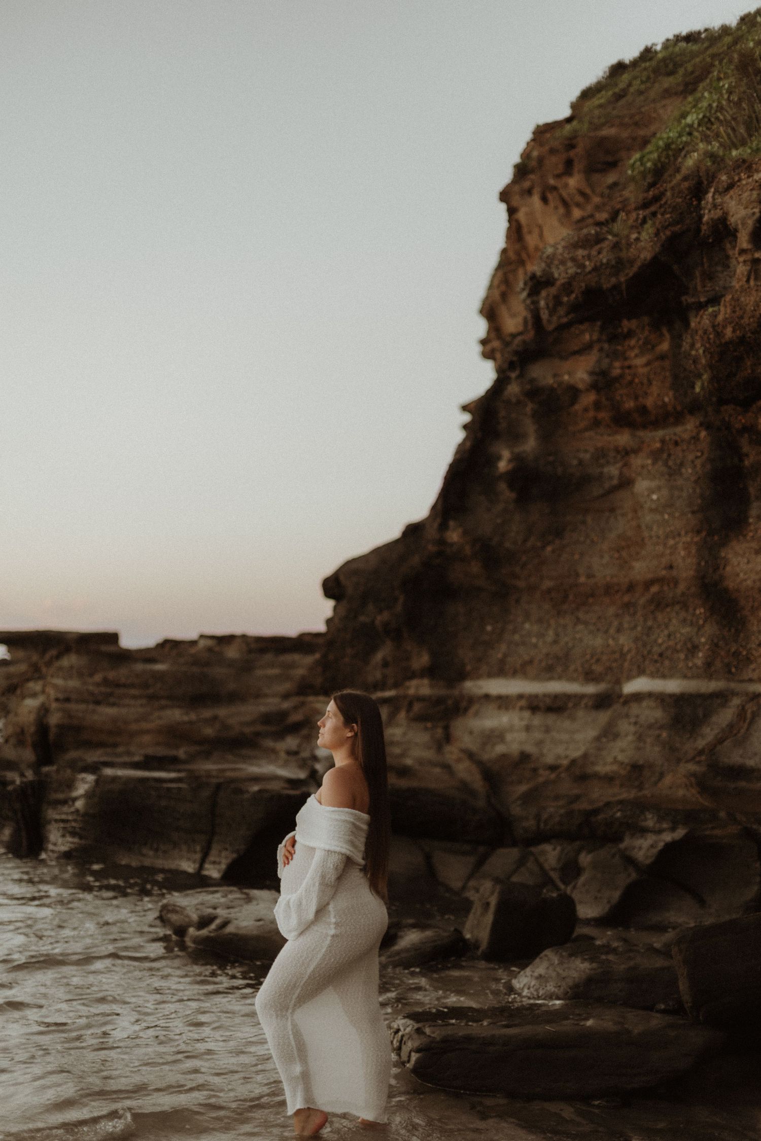 Pregnant woman in white dress standing by rocky coastal cliffs at dusk.