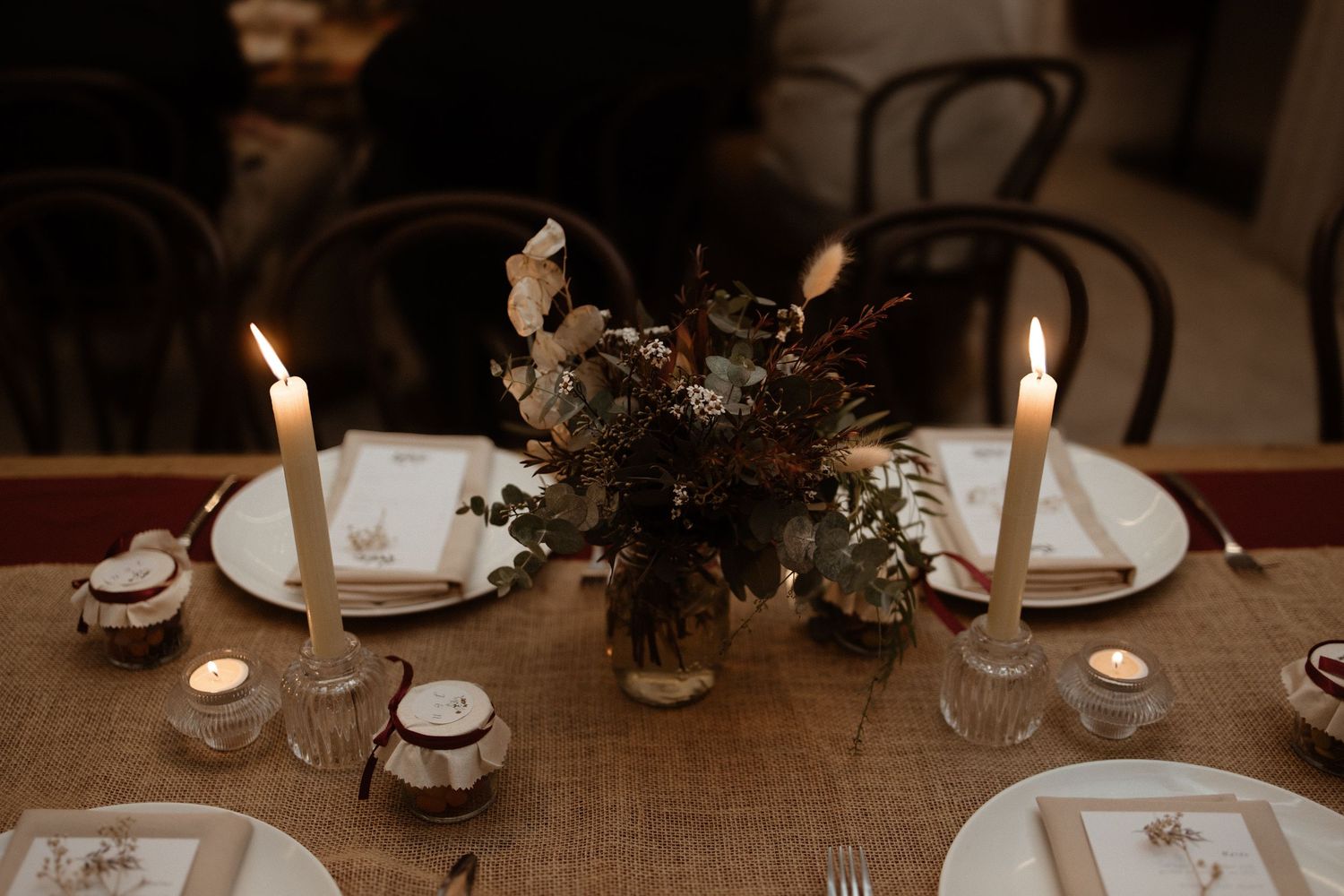 Elegant rustic wedding table centerpiece with dried florals, taper candles, and white place settings on burlap runner.