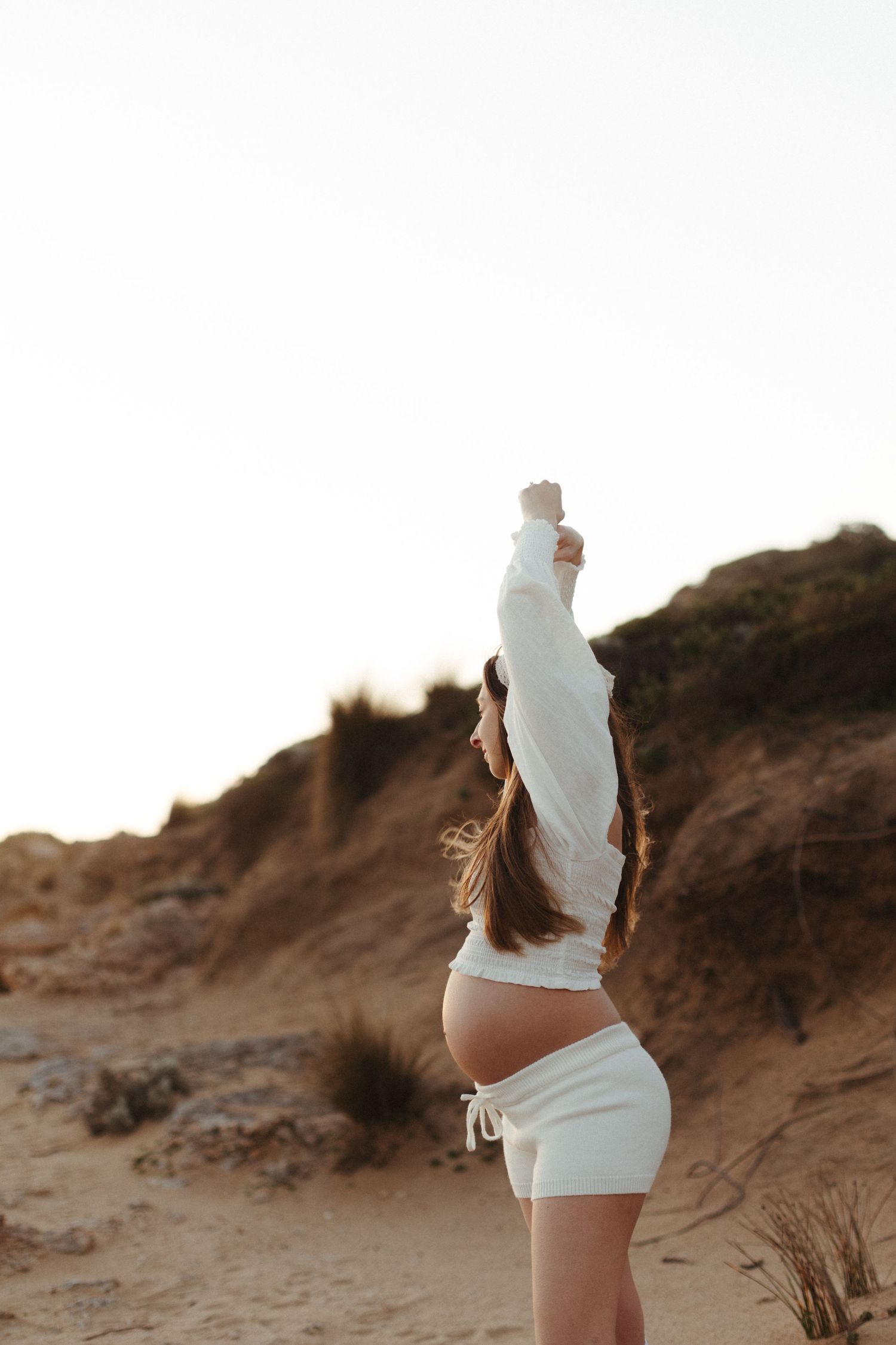 Pregnant woman in white outfit stretching arms overhead at golden hour on sandy desert dunes.