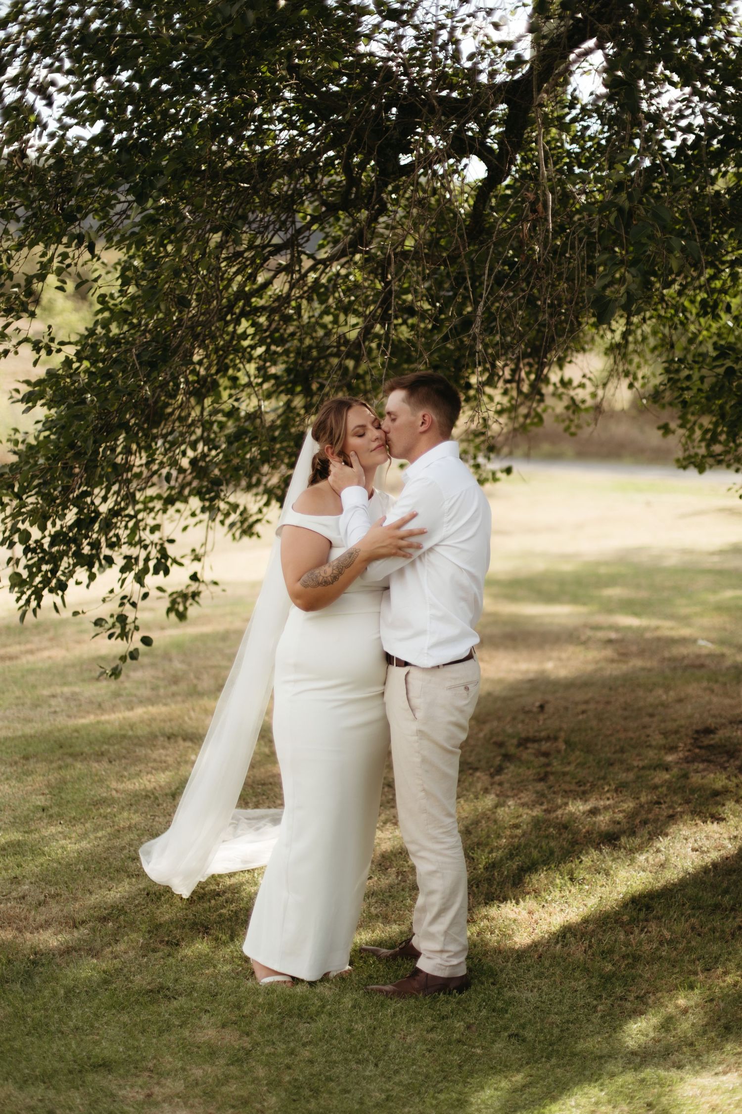 A bride and groom share a romantic kiss under a lush green tree on their wedding day outdoors.