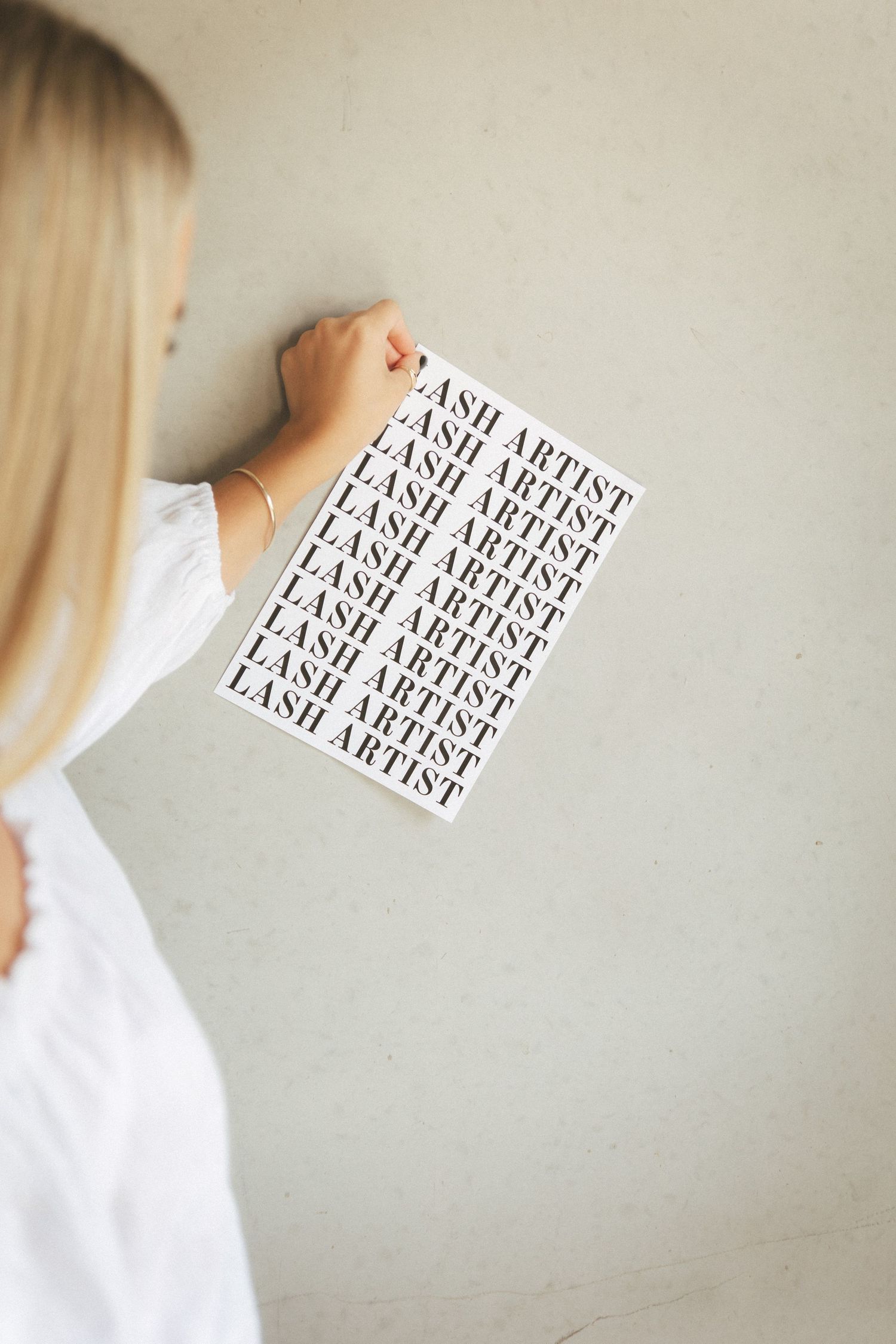 Blonde woman holding Lash Artist repeat-pattern print poster against light gray concrete wall.