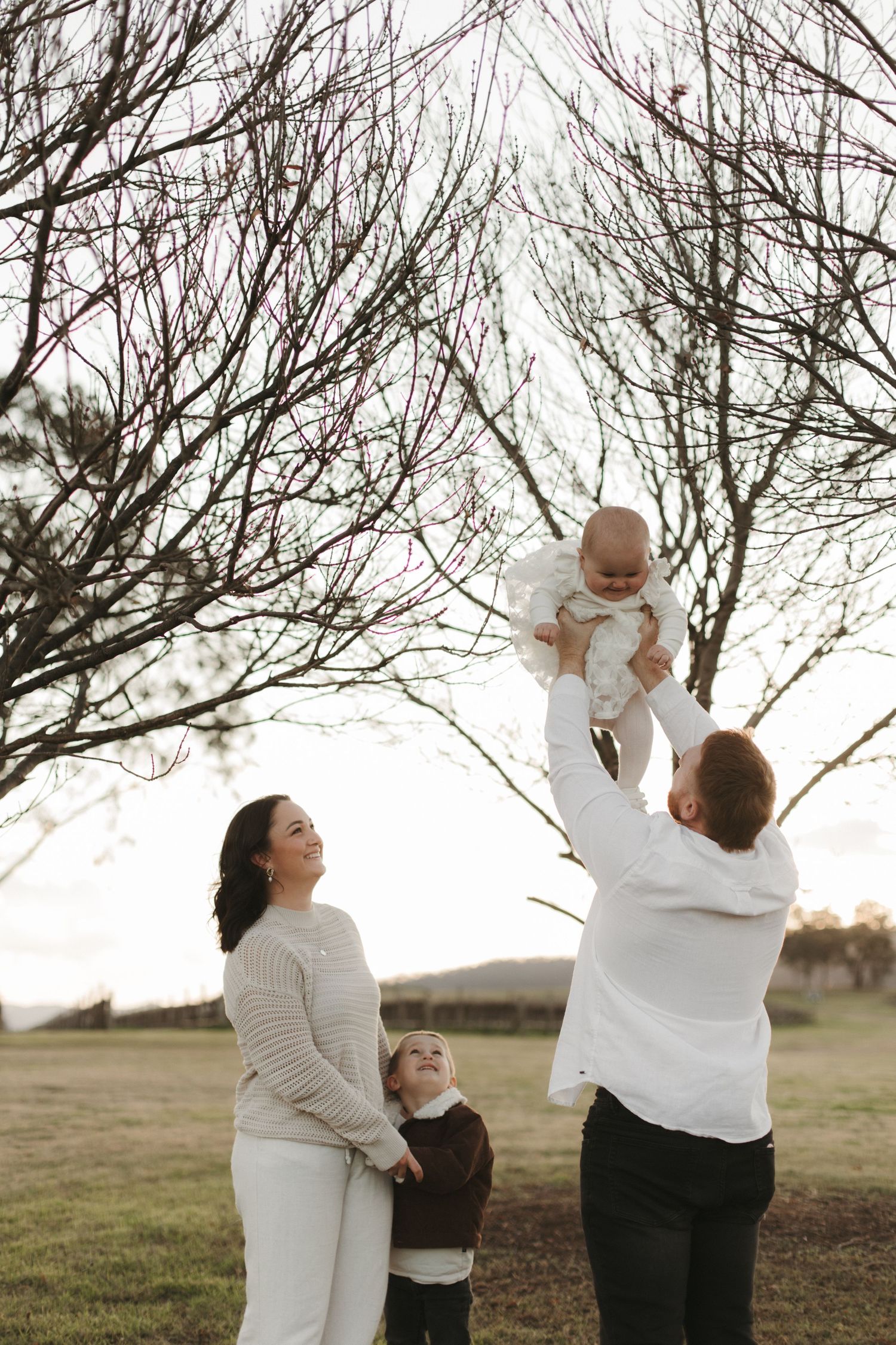 Family of four outdoors, father lifting baby in air near bare winter trees at golden hour.