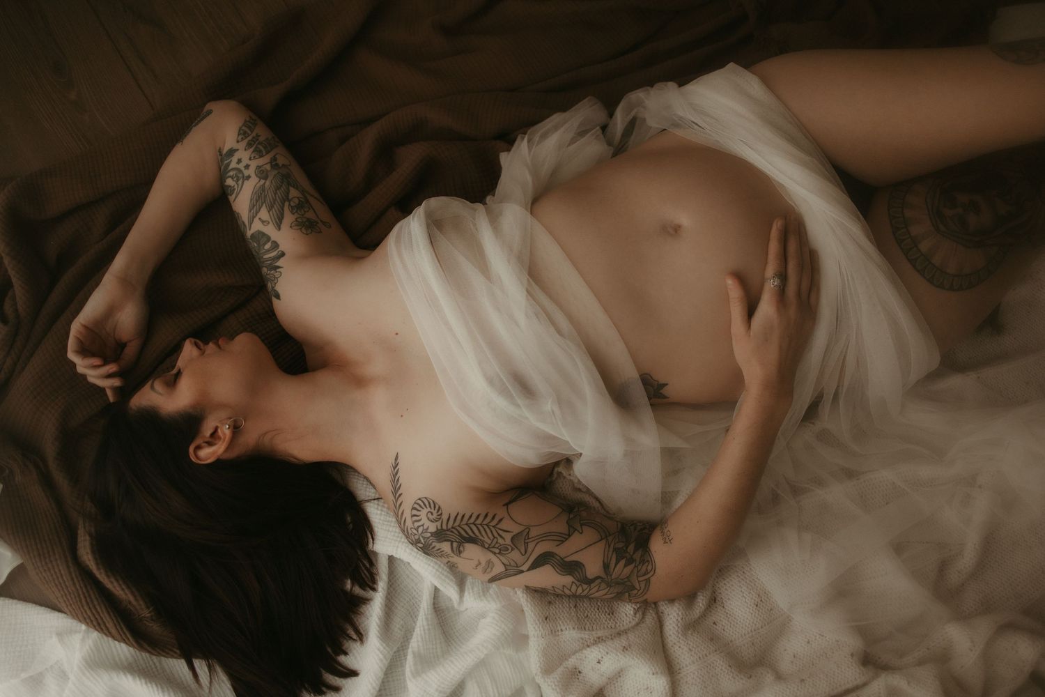 Tattooed pregnant woman lying on bed draped in white tulle fabric in moody indoor setting.