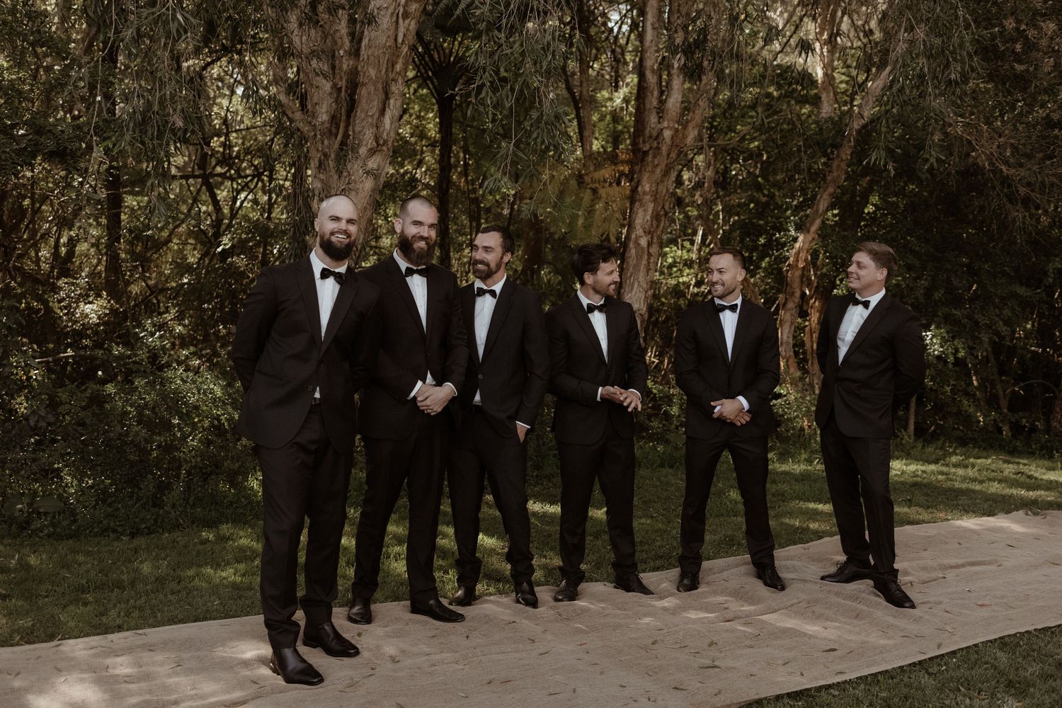 Six groomsmen in matching black tuxedos laughing together in a lush garden setting.