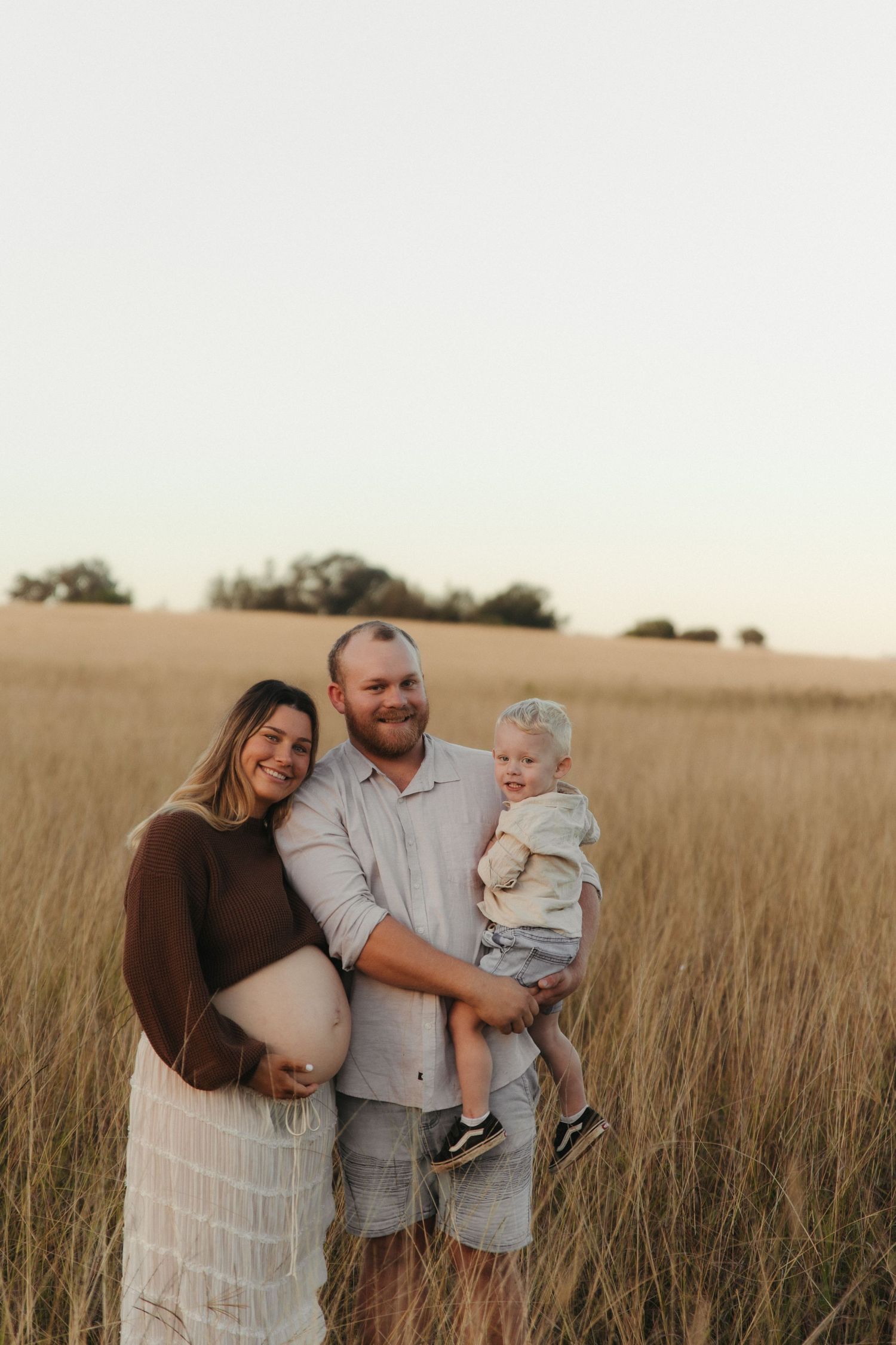 Pregnant woman and man holding toddler smiling in golden field at sunset during maternity photo session.
