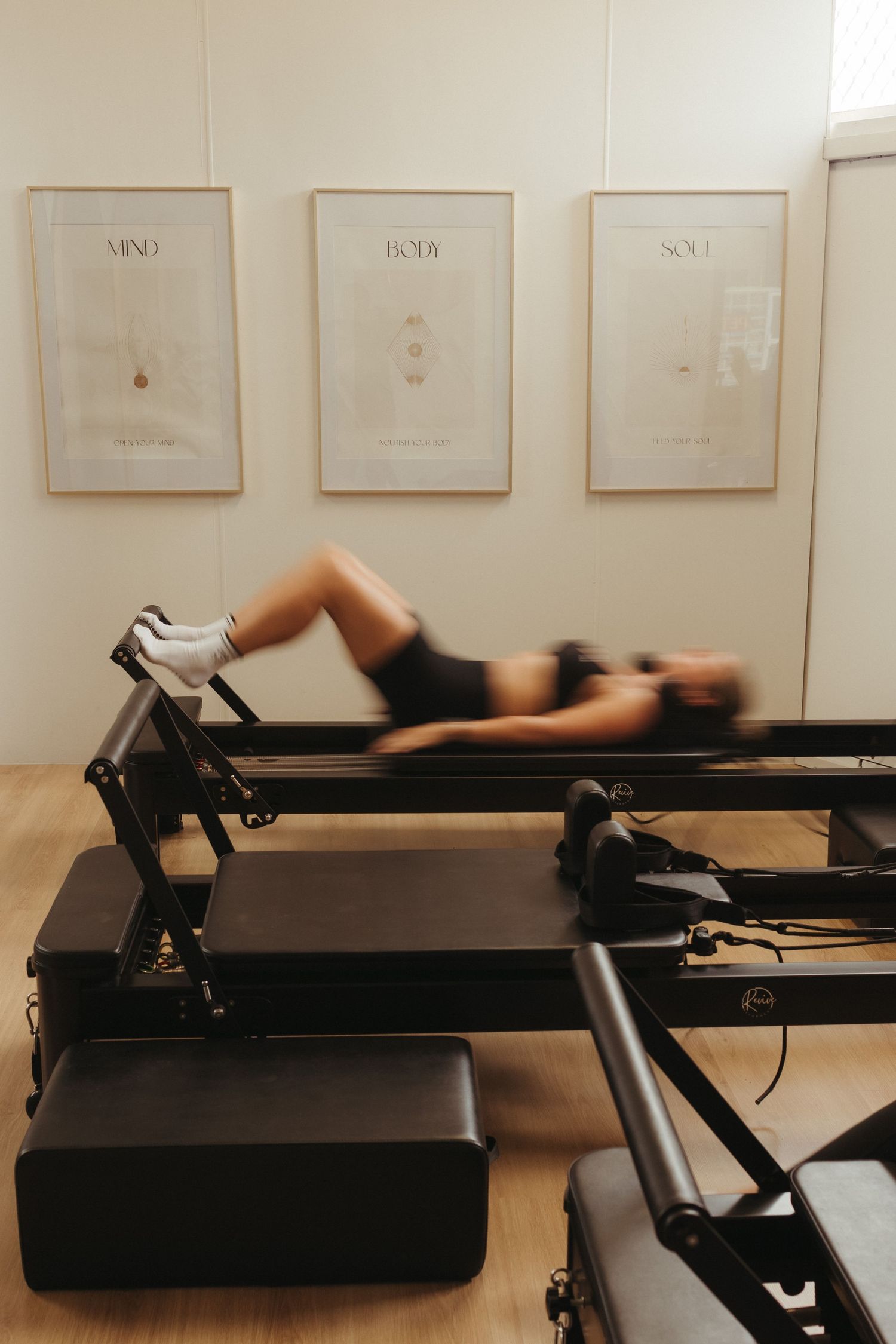 Person doing Pilates reformer exercise in bright studio with Mind Body Soul framed prints on wall.