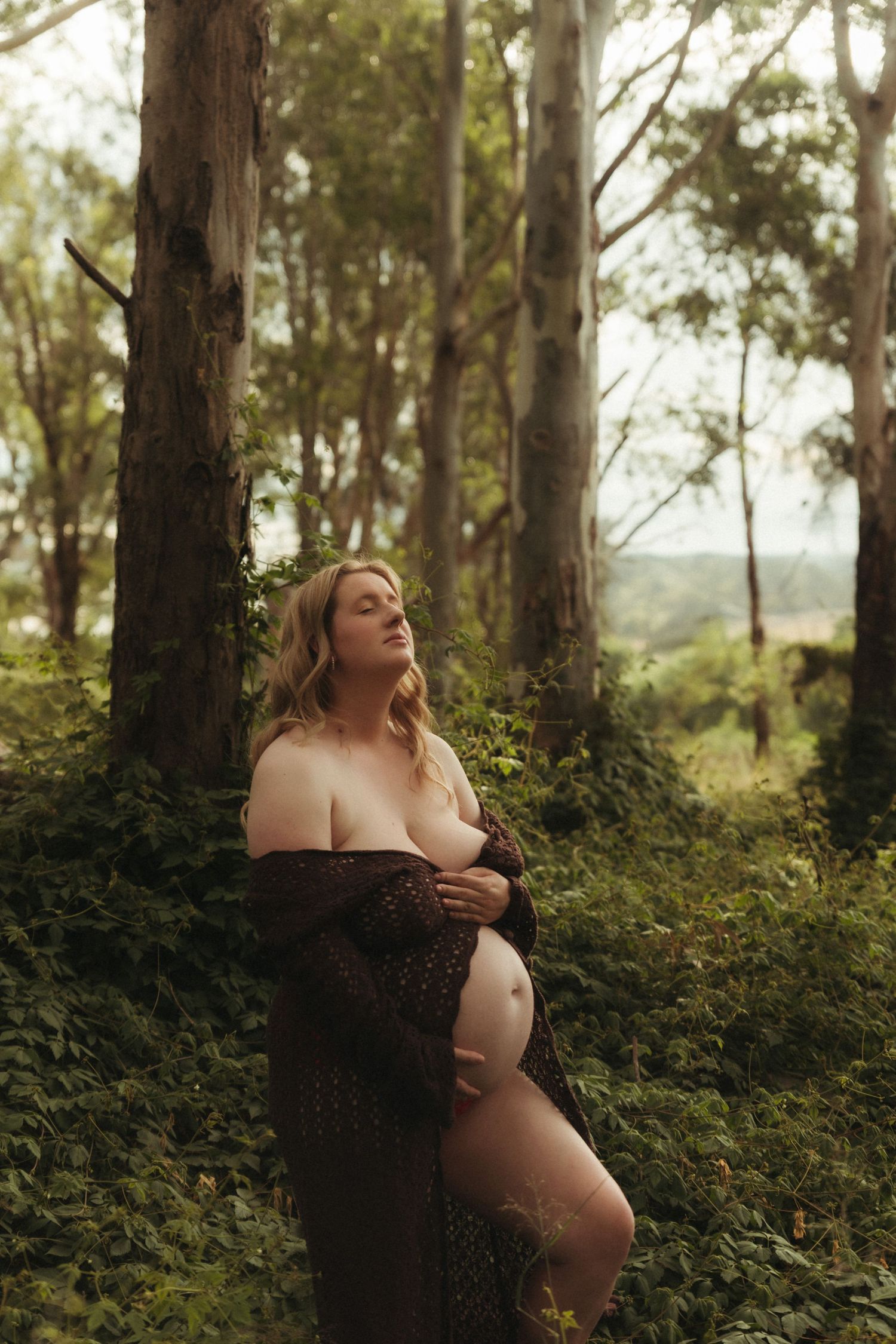 A pregnant woman in an open brown knit wrap leans against a tree in a lush green forest setting.
