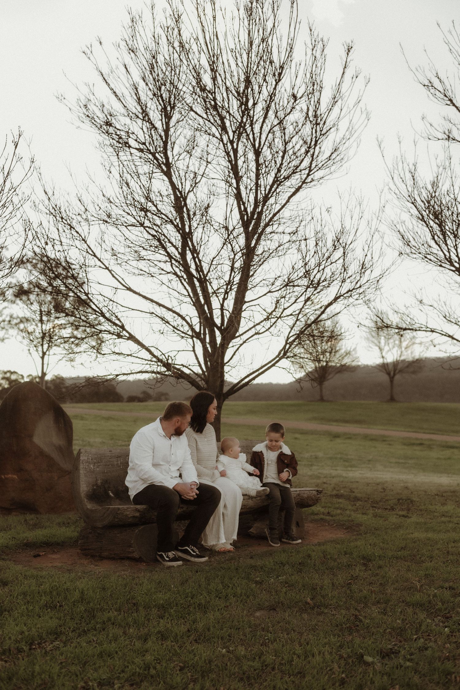 Family of four sitting together on a rustic log bench beneath bare trees in a green park.