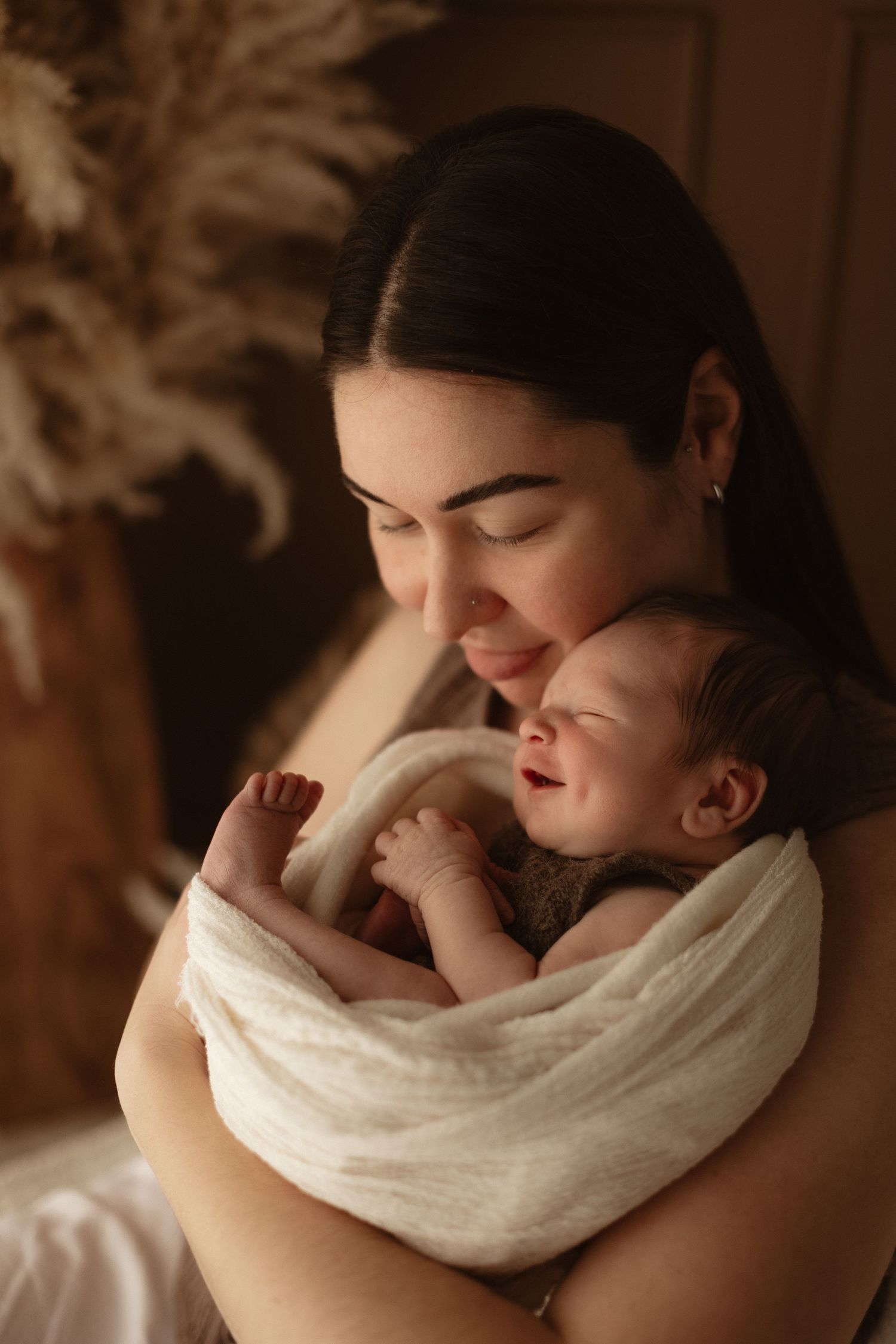 A mother tenderly holds her sleeping newborn baby wrapped in a white swaddle in warm studio lighting.