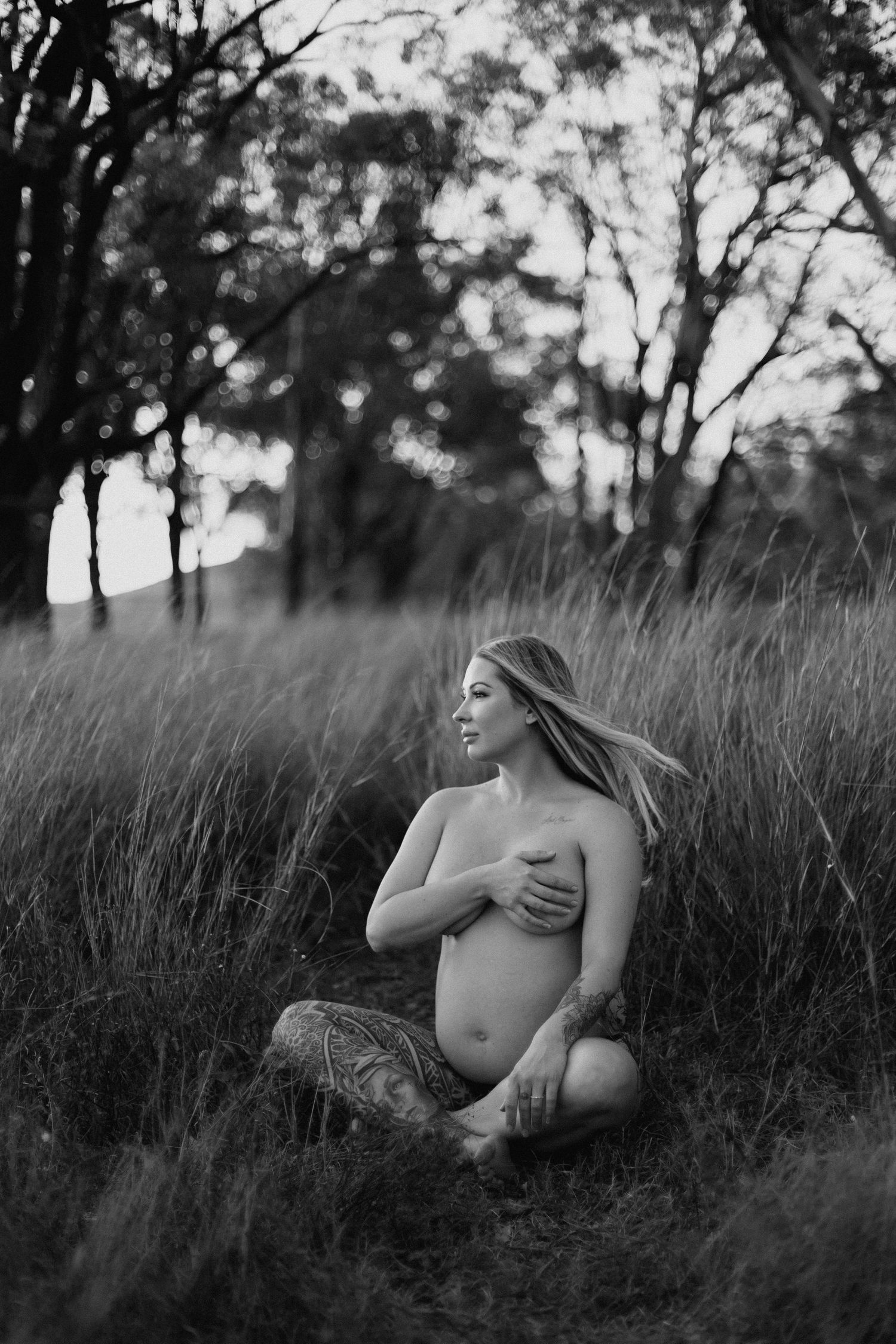 Black and white photo of topless pregnant woman sitting in tall grass field at sunset.