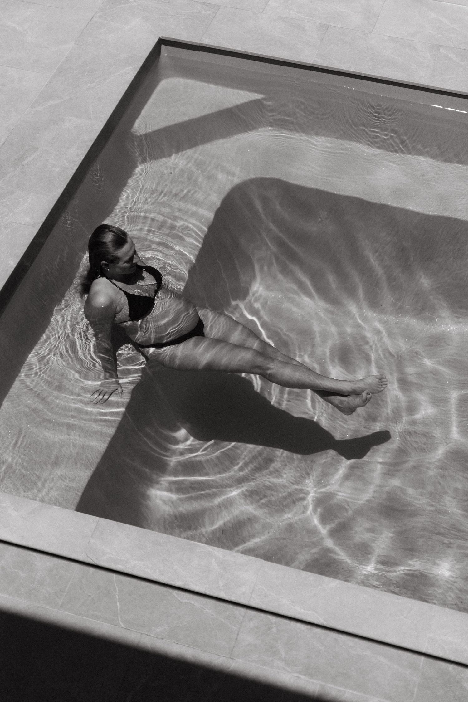 Black and white aerial view of a pregnant woman relaxing in a small square swimming pool.