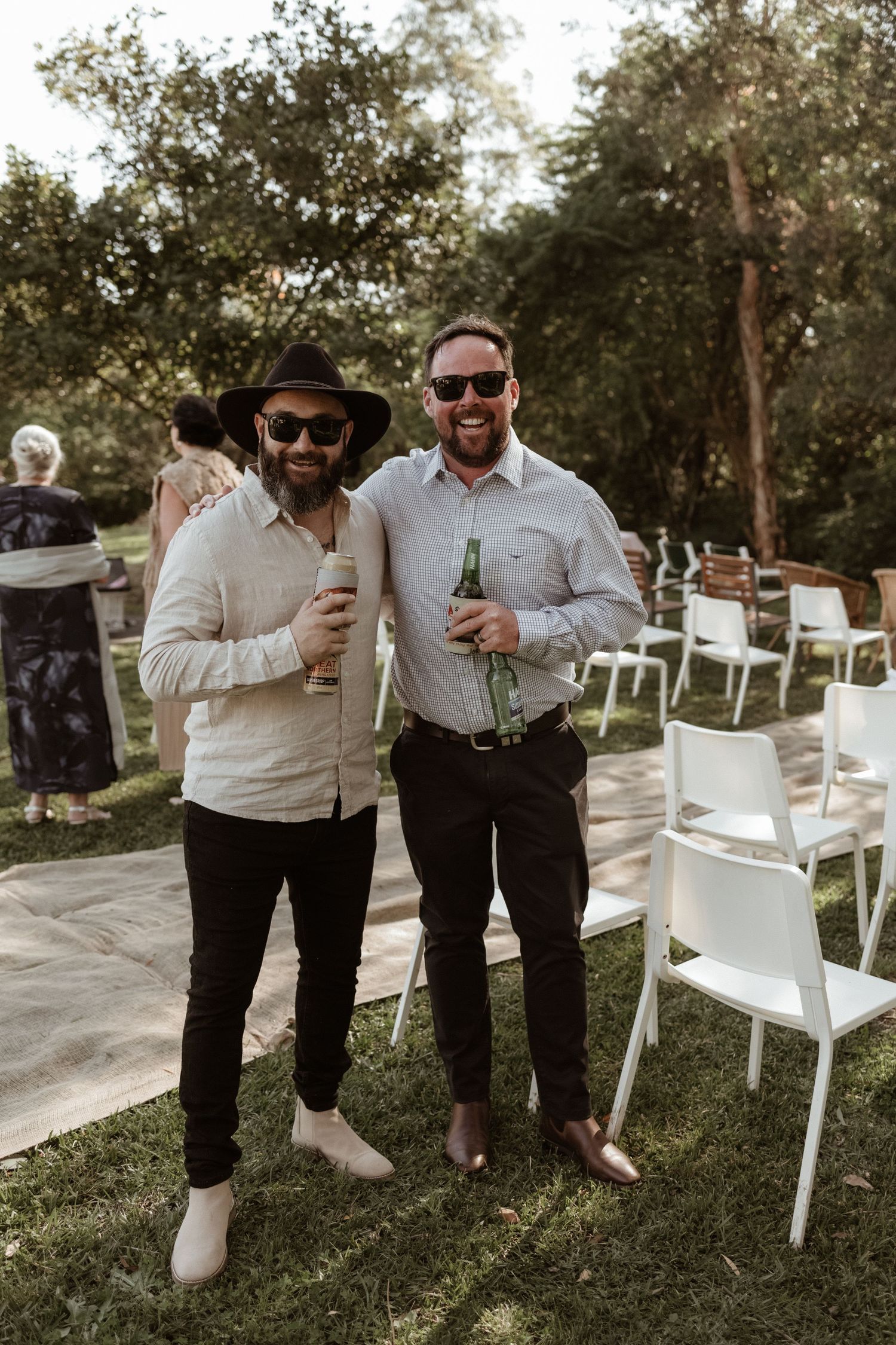 Two smiling male wedding guests holding beer bottles outdoors in sunglasses.