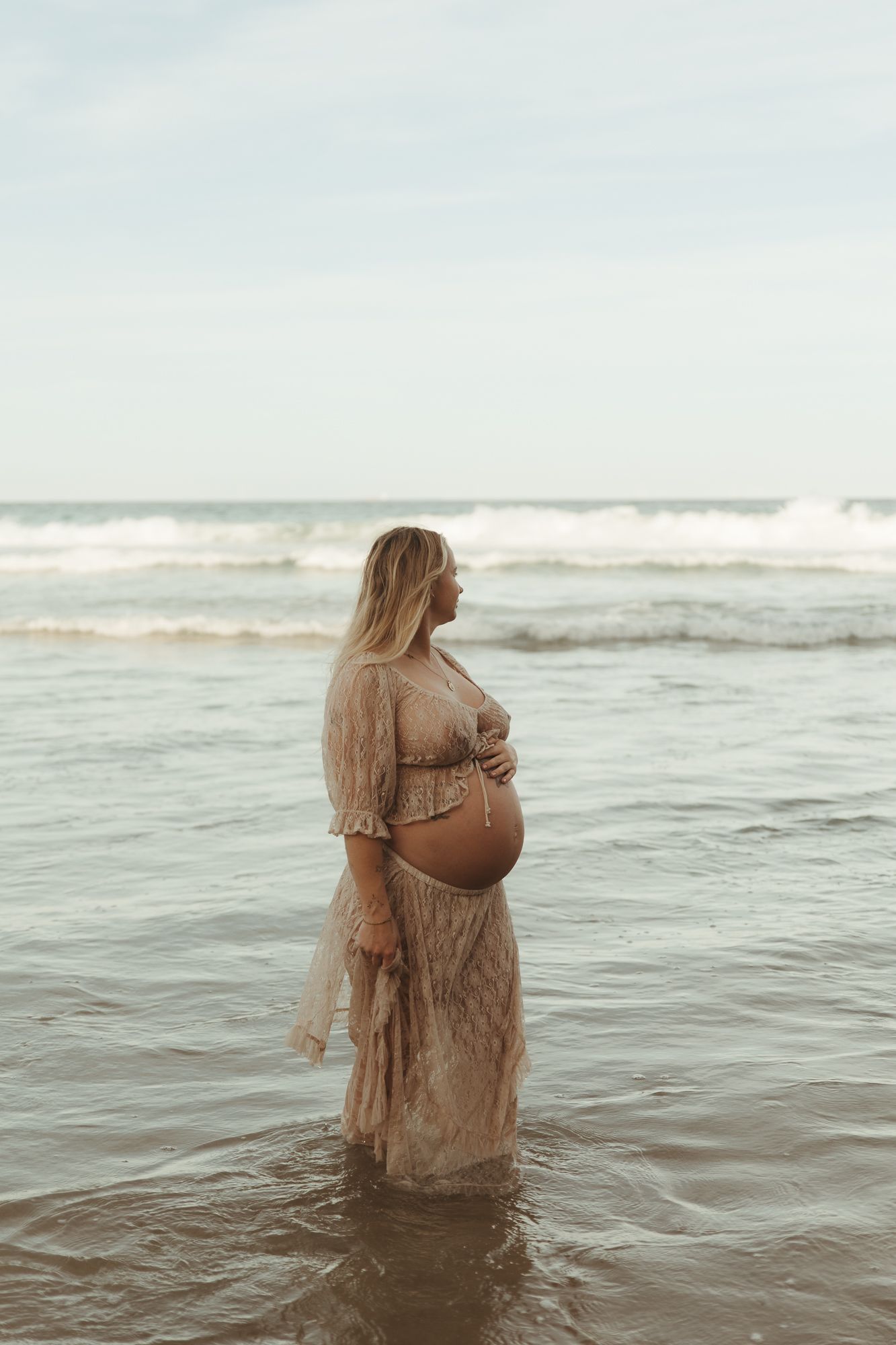 Pregnant woman in lace dress standing in ocean waves during maternity photoshoot at the beach.