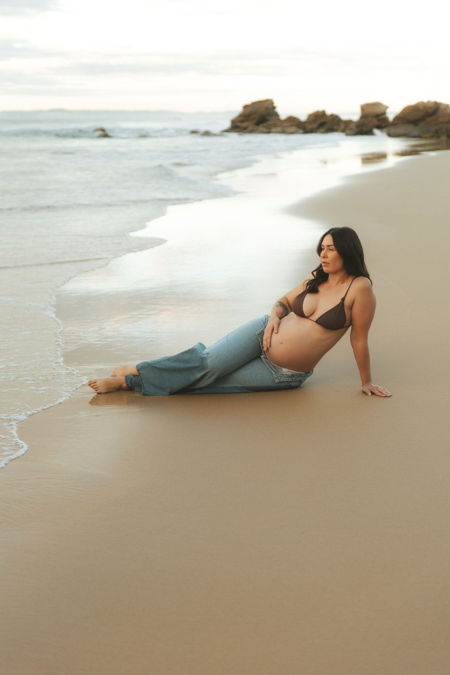 Pregnant woman in bikini top and jeans sitting on sandy beach shore with ocean waves, maternity photo session.