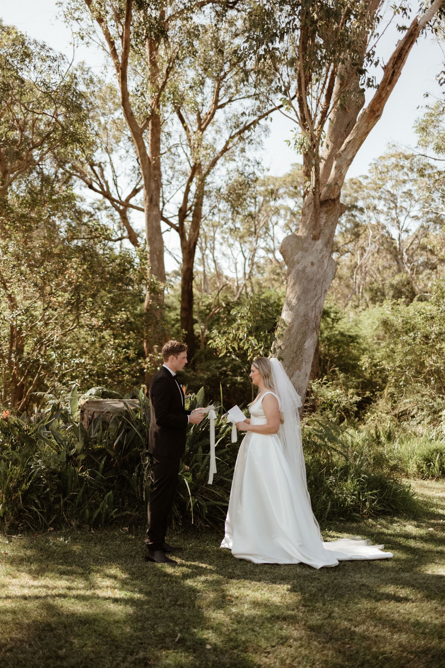 Bride and groom exchange vows outdoors in a lush garden setting, surrounded by tall eucalyptus trees.