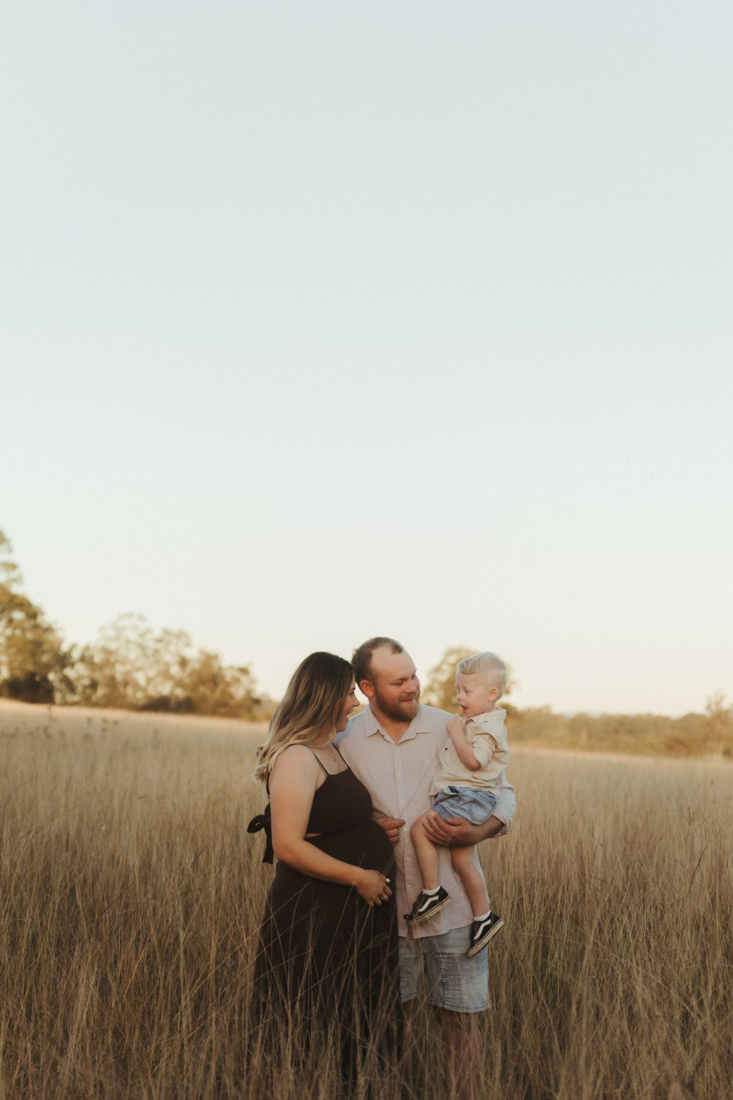 A couple stands in a golden field at sunset, smiling at their toddler held in the man's arms.