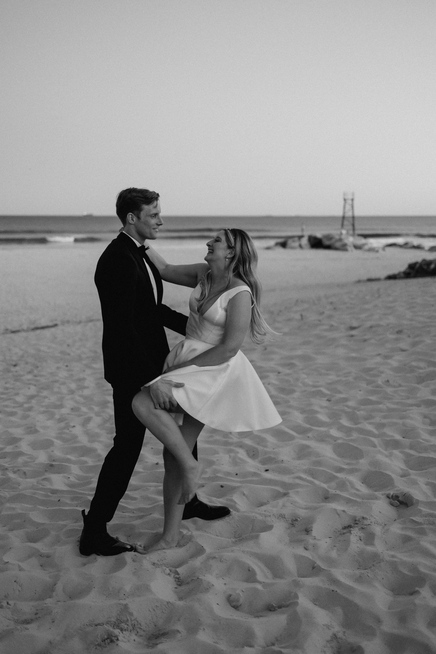Black and white photo of newlyweds dancing playfully on a sandy beach at dusk after wedding.