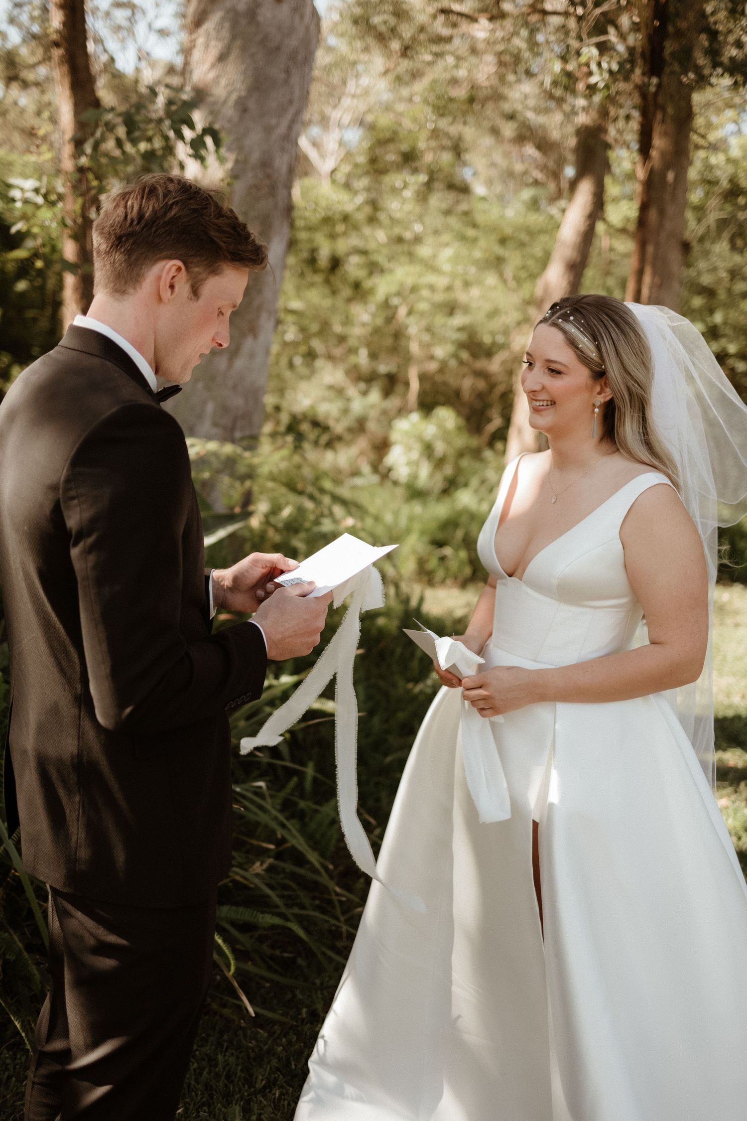 Smiling bride in white gown listens as groom reads wedding vows in a beautiful garden setting.