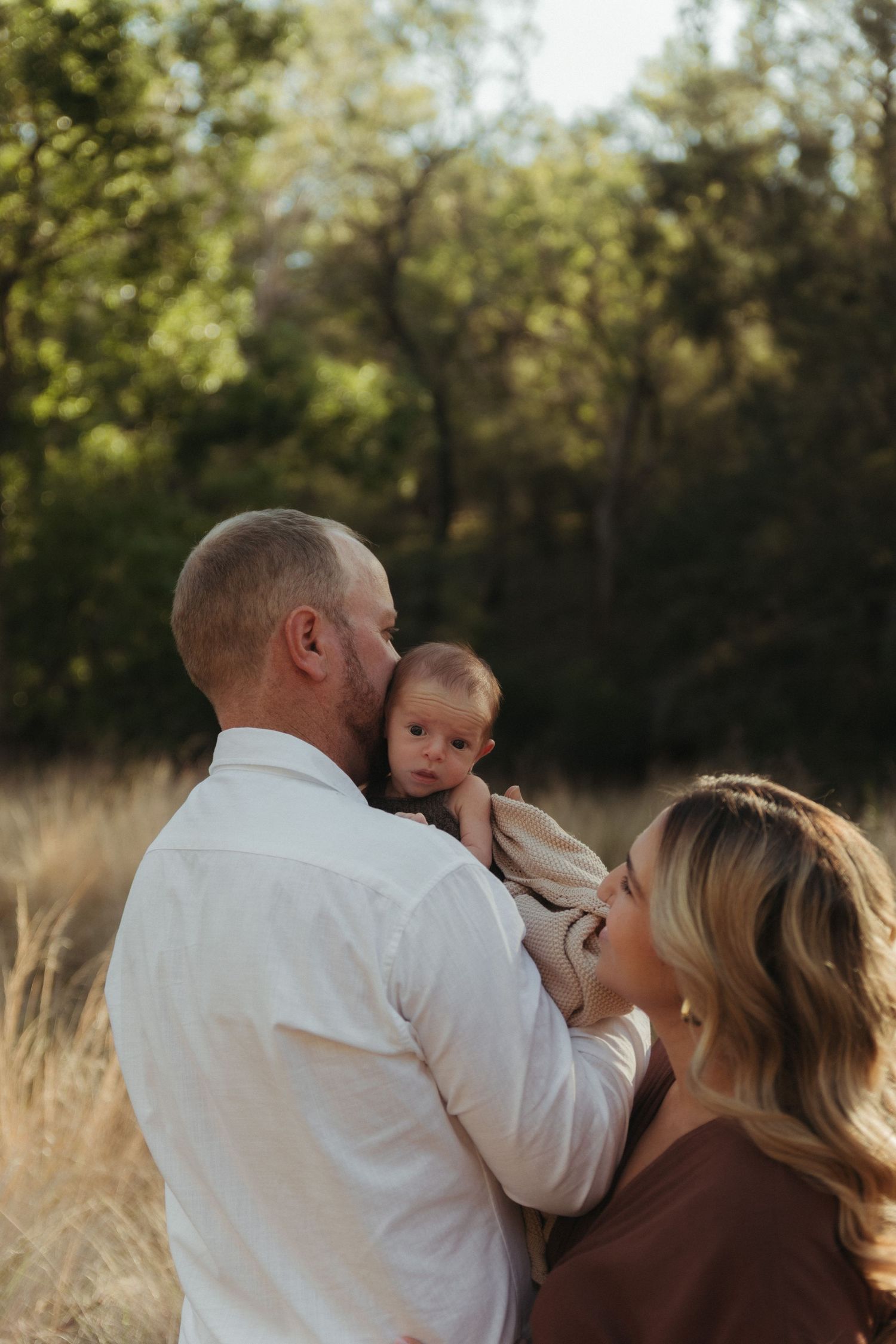 Parents sharing a tender moment with their alert newborn baby in a sunlit outdoor field setting.
